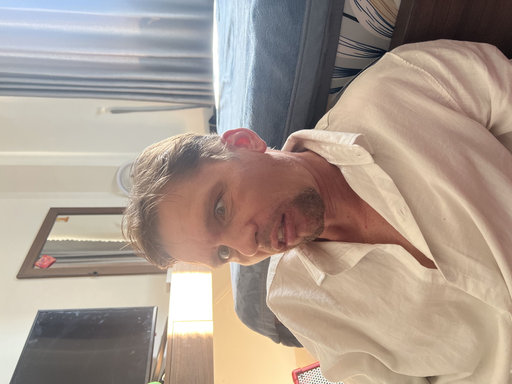 A man is lying down in a room, possibly a bedroom. He is wearing a white linen shirt and appears to be looking at the camera with a curious or surprised expression. The room is lit by natural light, suggesting it is daytime. In the background, a bed with a blue cover is visible, as is a mirror on the wall reflecting a string of lights and a red object. A television screen is also visible in the lower left corner of the image. The overall impression is of a casual, indoor setting, possibly in a residential or hotel room in Da Nang, Vietnam. There is no visible text in the image.