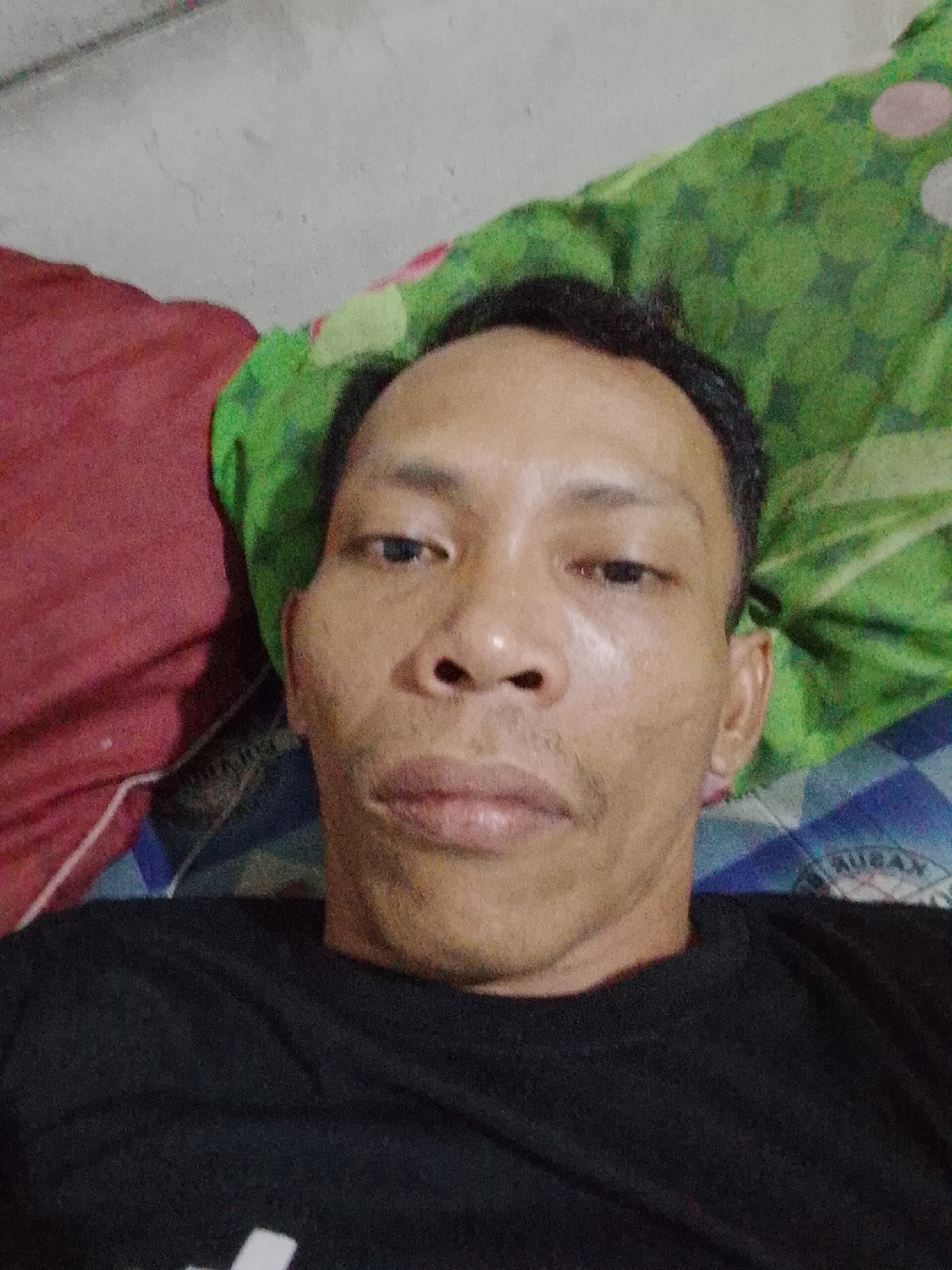 A man is lying down, looking up at the camera. He is wearing a black shirt. The background consists of a red pillow on the left and a green patterned blanket on the right. The setting is likely indoors. There is no specific information to definitively place the image in Bukit Ketok, Indonesia; that is an assumption based on prior context.