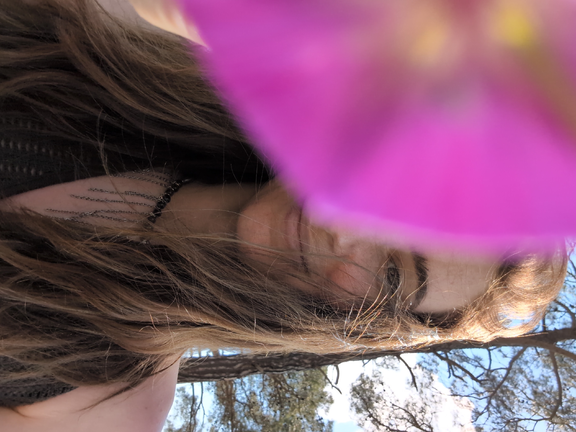 In a close-up, outdoor photograph, the right side of a young woman's face is visible, partially obscured by a large, out-of-focus pink petal. She has fair skin and long, light brown hair that is windswept. She appears to be looking slightly to her left. Her left eye, eyebrows, and the bridge of her nose are visible. She is wearing a dark, intricate necklace with multiple dangling chains. Her right shoulder and upper arm are also visible, and she is wearing a dark, possibly knitted, top. Behind her, a pine tree with visible branches and a bright blue sky with some white clouds suggest a natural, outdoor setting. There is no indication of the location being in Divnomorskoye, Russia.