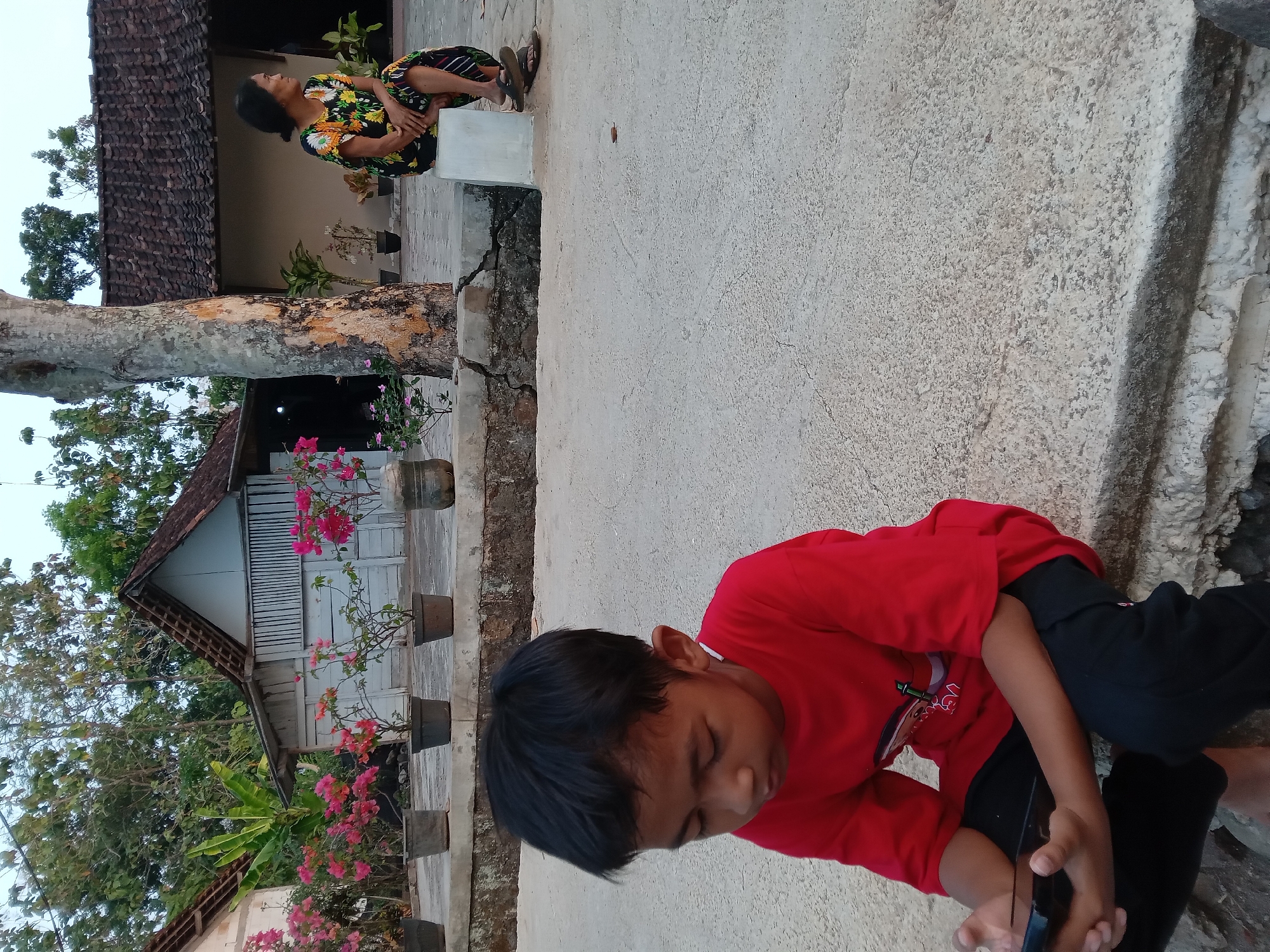 In Kedunggudel, Indonesia, a young boy with dark hair, wearing a red t-shirt and black pants, sits on a concrete surface, engrossed in his mobile phone. To his right, a woman with dark hair, dressed in a colorful floral print dress, sits on a white block, looking away from the camera. Behind them, a large tree trunk stands prominently. The background features traditional Indonesian houses with tiled roofs and several potted plants, including vibrant pink bougainvillea. The overall environment suggests a rural or semi-rural setting with a sense of everyday life.