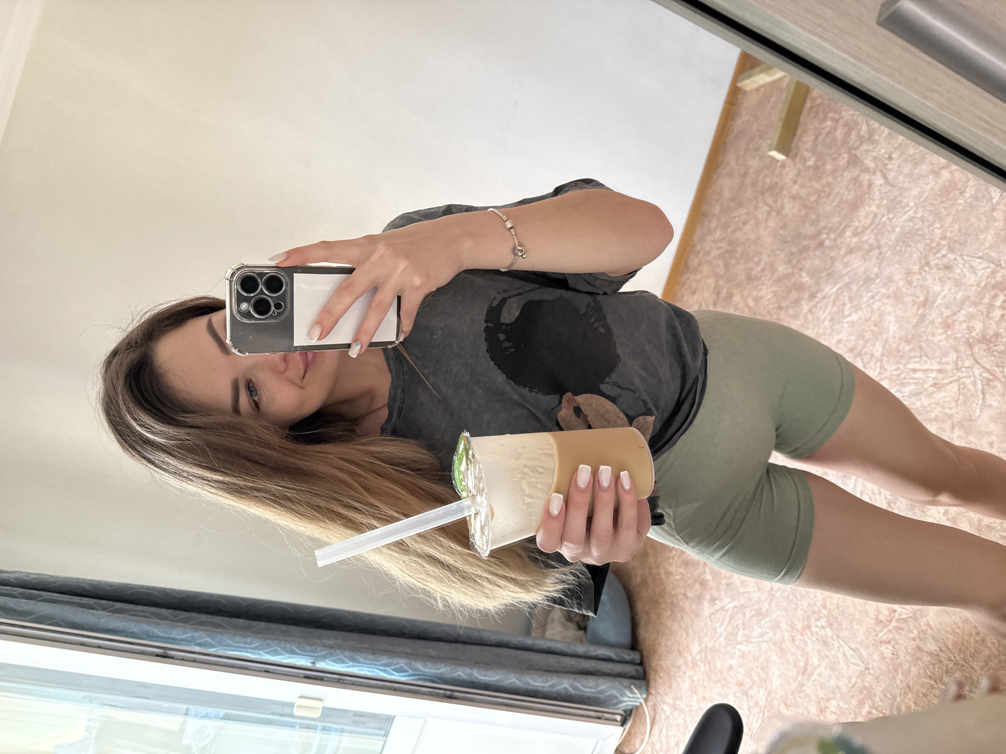 A woman is captured in a mirror selfie, holding a smartphone in her left hand and a beverage in her right. The image is vertically oriented, showing the woman's torso, legs, and part of her face reflected in the mirror.
The woman has long, straight, light brown hair that falls over her shoulders. Her face is partially visible in the phone's reflection, showing her right eye, part of her nose, and a neutral expression. She is wearing a dark grey t-shirt with a faded, indistinct graphic design on the chest, and light olive green biker shorts. A silver charm bracelet is on her left wrist. Her fingernails are neatly manicured with a light-colored polish, possibly a French tip style, and a small blue accent on one finger.
She holds a clear plastic cup containing a layered drink, which appears to be bubble tea or a similar beverage with a creamy top layer and a darker brown liquid below. A wide, clear straw is inserted through the sealed lid of the cup. The smartphone, an iPhone model with a prominent triple-lens camera system, is encased in a clear protective cover.
The background, visible as a reflection in the mirror, suggests an indoor setting. It includes a light-colored wall or ceiling, a textured light brown or beige carpet or flooring, and a window with blue horizontal blinds or curtains in the lower left section. Part of a door frame or cabinet is visible in the upper right of the reflection. The scene conveys a casual, everyday moment.