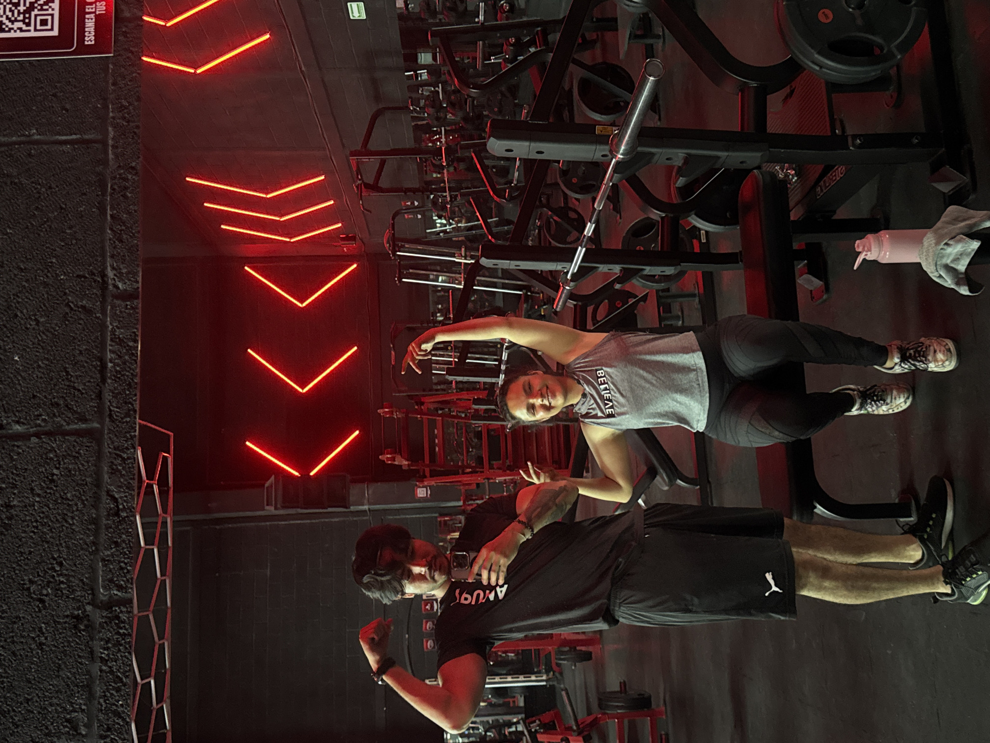 The image depicts two people in a gym setting, likely in the city of Puebla, Mexico, based on possible context clues that are not visible in the image. A man in a black t-shirt and shorts is flexing his arm while holding a phone as if taking a selfie. A woman beside him is gesturing with her hands. The gym has workout equipment, including weight racks and machines. The walls are dark, and red neon lights are mounted. A water bottle and towel are placed on a weight bench.