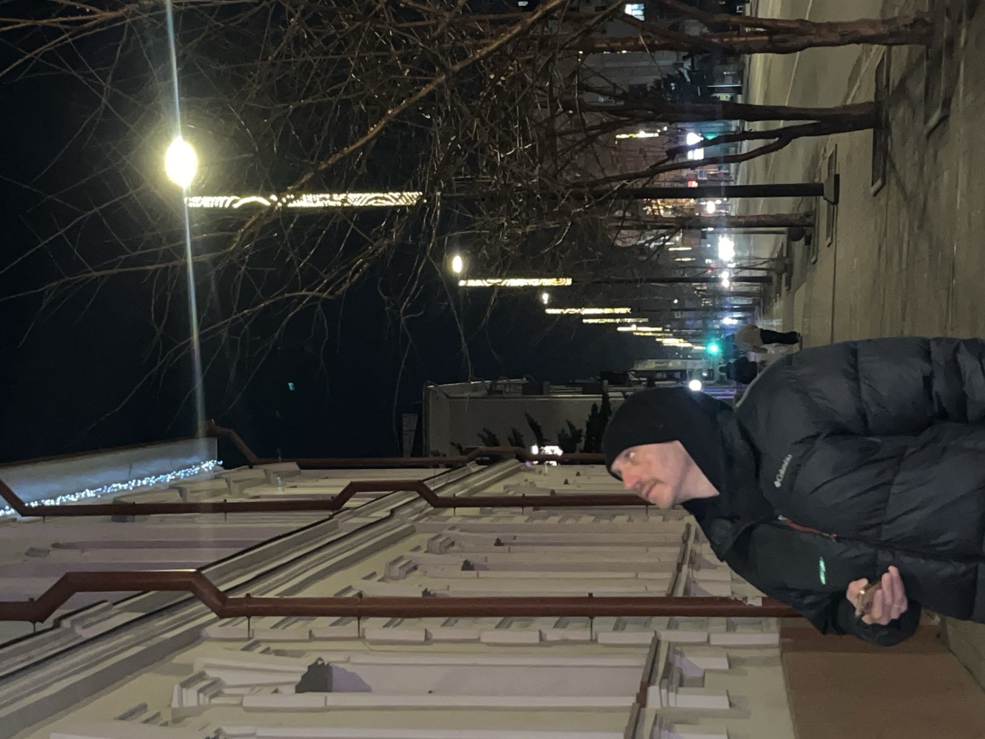 At night, in a city (likely Krasnoyarsk, Russia), a man in a black hooded jacket and black beanie hat stands on a sidewalk next to a building. The building has a light-colored facade with several horizontal pipes running along it. There are lights strung along the top of the building. Above the building are trees with bare branches. Streetlights are visible in the background, illuminating the street, and there are other pedestrians further down the sidewalk.