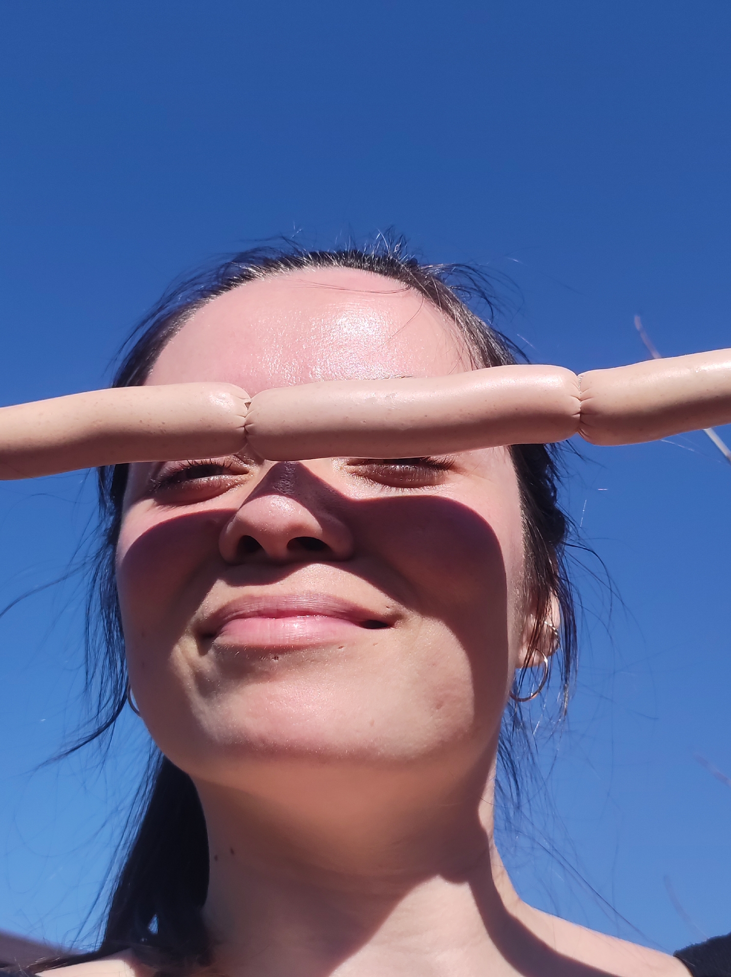 A woman is holding two sausages against her eyes. She is looking at the camera with a slight smile. The background is a clear blue sky. The location is not discernible.