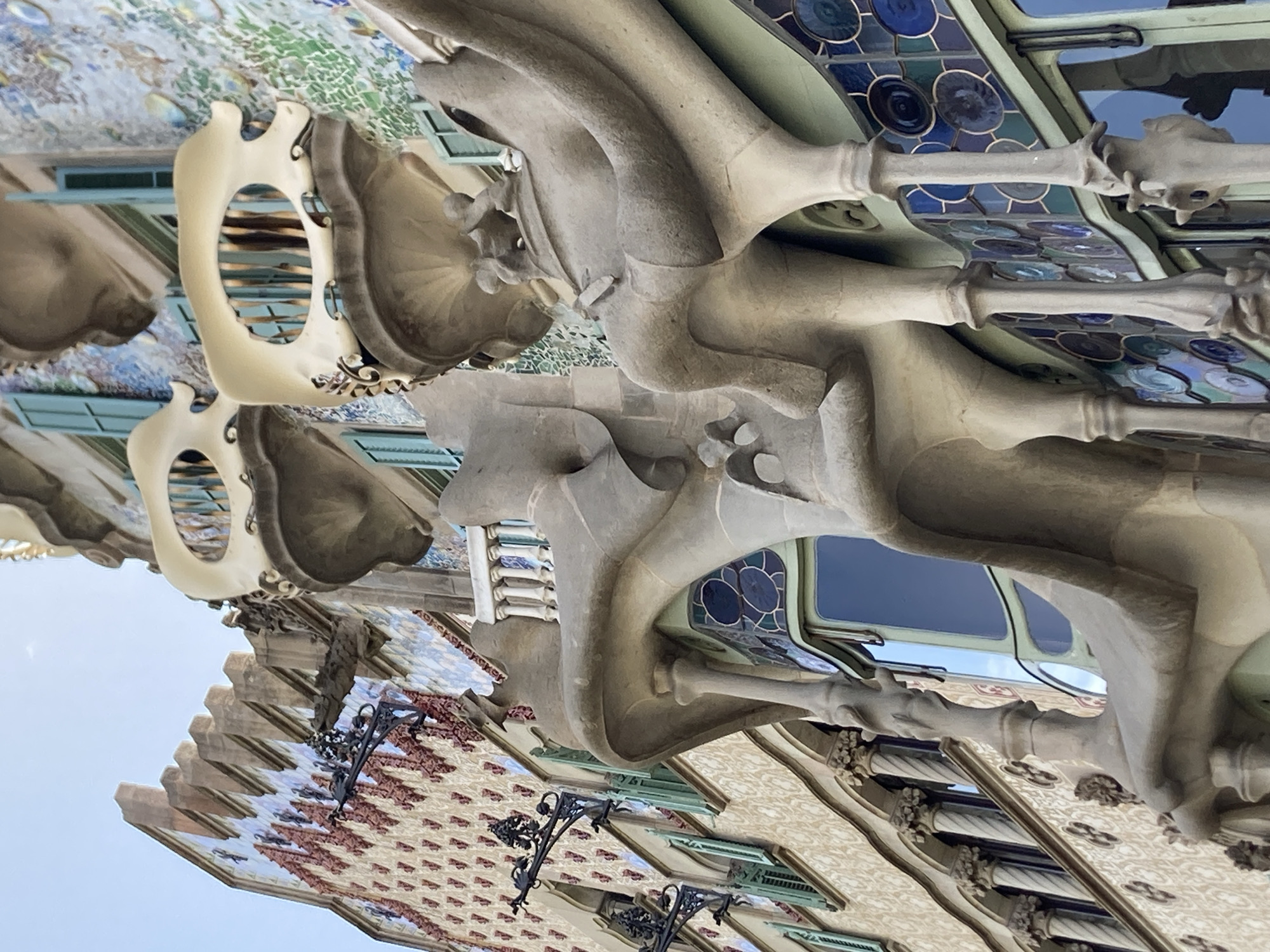 The image is a close-up, low-angle shot of the facade of Casa Batlló, a building located on Passeig de Gràcia in the Eixample district of Barcelona, Spain. The architecture is characterized by its organic, undulating forms and vibrant, mosaic-like exterior. In the foreground, sculpted stone elements, resembling bones or branches, form balconies and support structures. These elements are intricately detailed and appear to be made of a light-colored stone. Behind these structures, the building's facade is covered in a mosaic of colorful tiles, predominantly in shades of blue, green, and white, creating a shimmering, almost iridescent effect. Some of the tiles are circular, while others form more complex patterns. Several windows with turquoise shutters are visible, adding to the building's distinctive color palette. Decorative wrought-iron elements, possibly lamp holders, are attached to the upper portions of the facade. The sky, a clear pale blue, is visible in the upper left corner. The image focuses on the architectural details, highlighting the unique and artistic style of the building, a masterpiece of Antoni Gaudí's work.