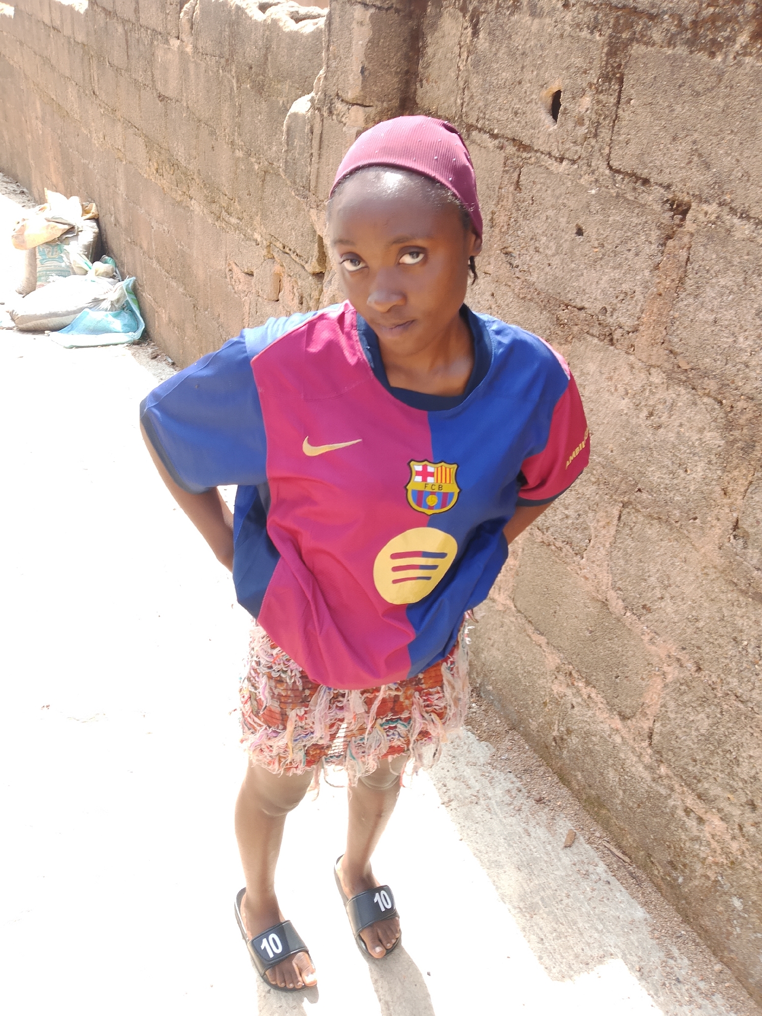 A young woman stands on a paved surface, her body angled away from the camera. She wears a pink and blue short-sleeved shirt with a Nike logo and a Barcelona Football Club crest, a patterned skirt, and black slide sandals with the number "10" on them. Her dark hair is covered by a burgundy headscarf. She is positioned in front of a rough, concrete wall, which appears to be part of a building or enclosure. Bags of what appear to be cement or construction materials are stacked to the left of the woman and the wall. The scene is brightly lit, suggesting daylight. The style of the wall and the presence of construction materials suggest an outdoor location, possibly a construction site or an undeveloped area within a city. Considering the urban environment and construction elements, the location is consistent with an outdoor setting in Jos, Nigeria.