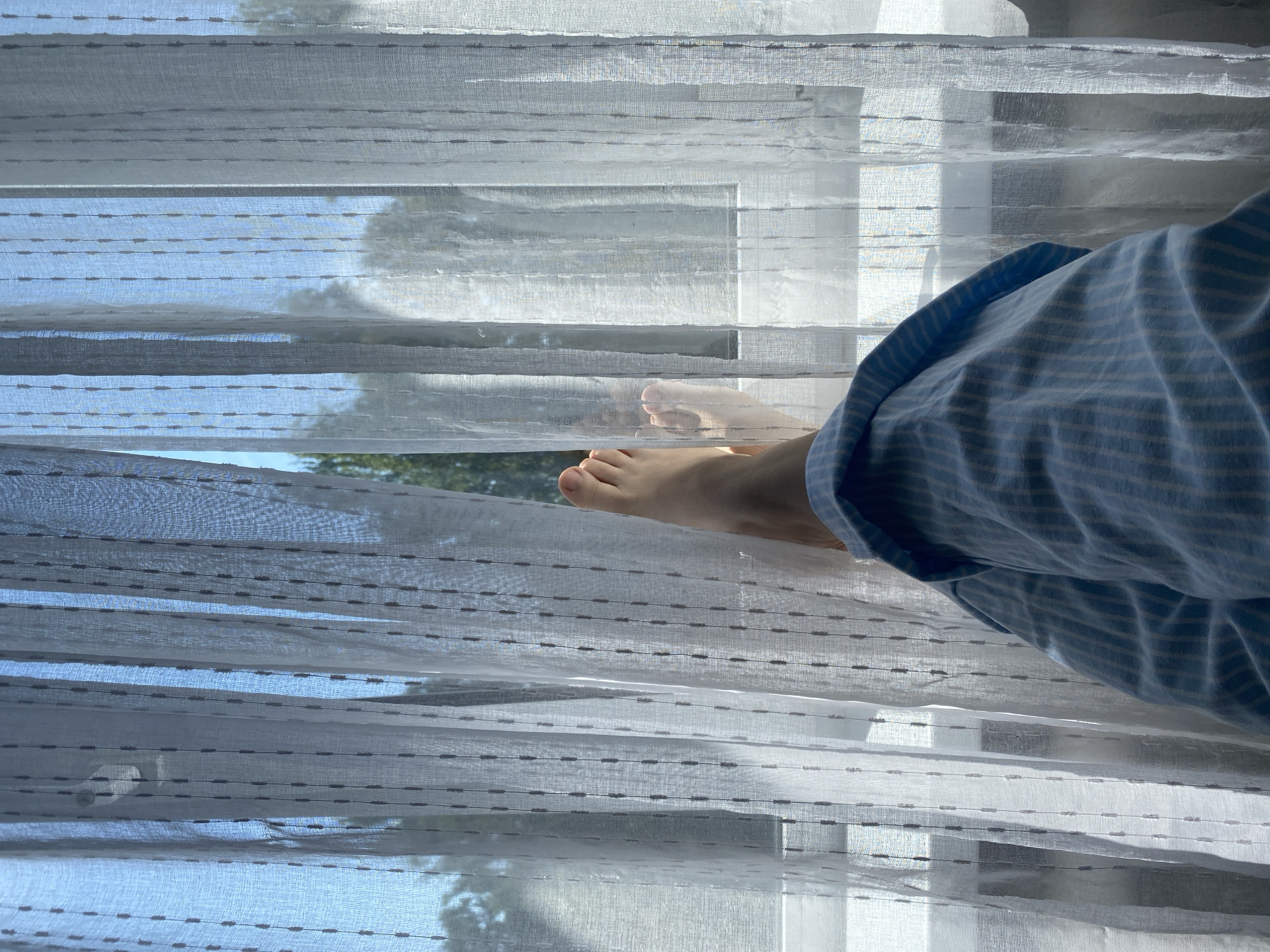 The image shows a person's legs and feet from a first-person perspective, resting in front of a window with sheer white curtains. The person is wearing light blue and white striped pajama pants. The curtains are slightly parted, revealing a view of trees and a bright blue sky outside. Sunlight streams through the curtains, creating a soft, diffused light. The scene appears to be indoors, possibly in a bedroom or living room. There is no indication of the specific location within the city of Chaikovsky, Russia.