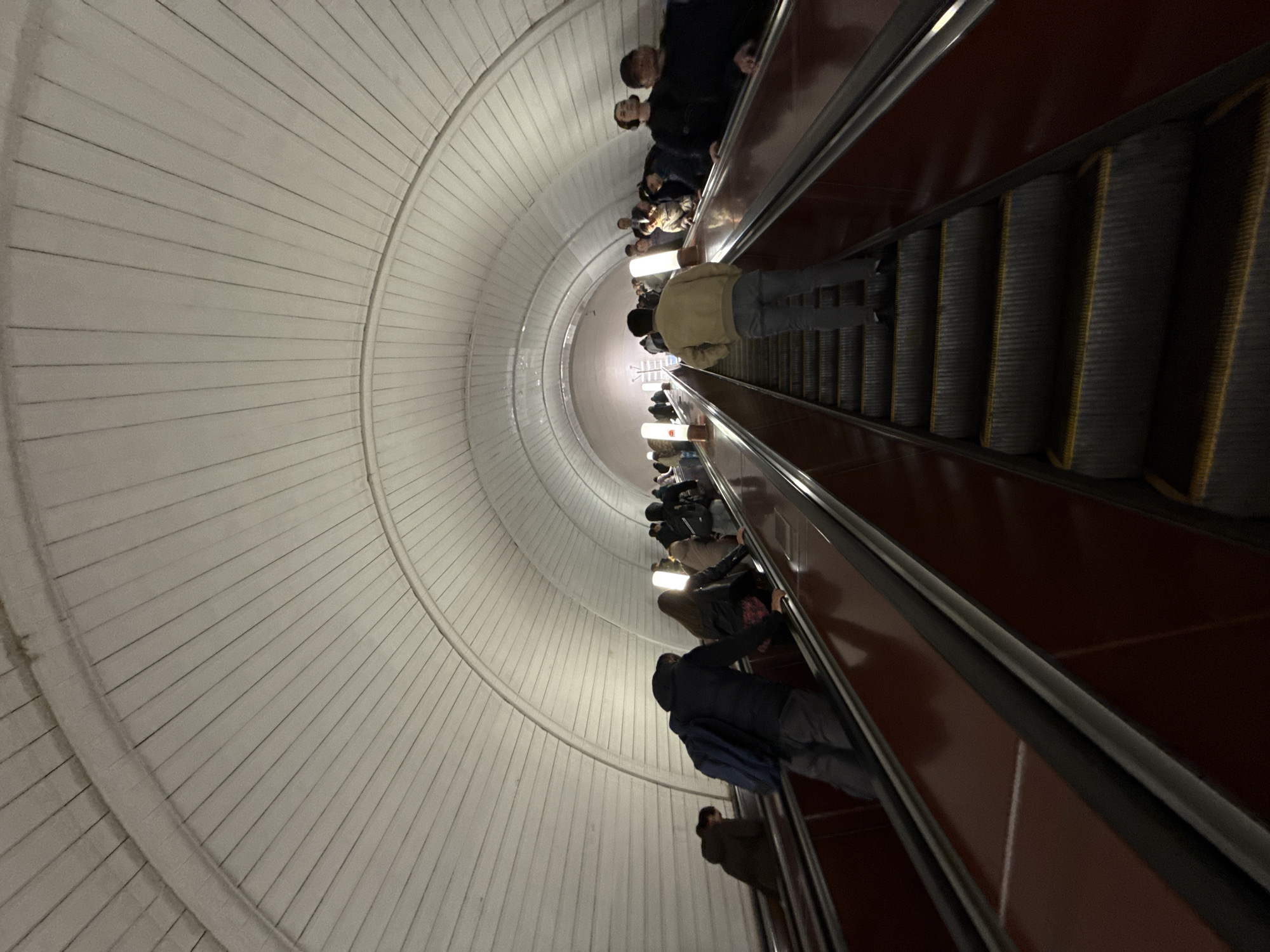 The image depicts people riding down a long escalator in a Moscow metro station, Russia. The escalator is enclosed by a tunnel-like structure with white, radiating lines on the walls. Passengers are descending on the right side of the frame, with a line of people going down the escalator. The lighting in the tunnel is bright and diffuse.