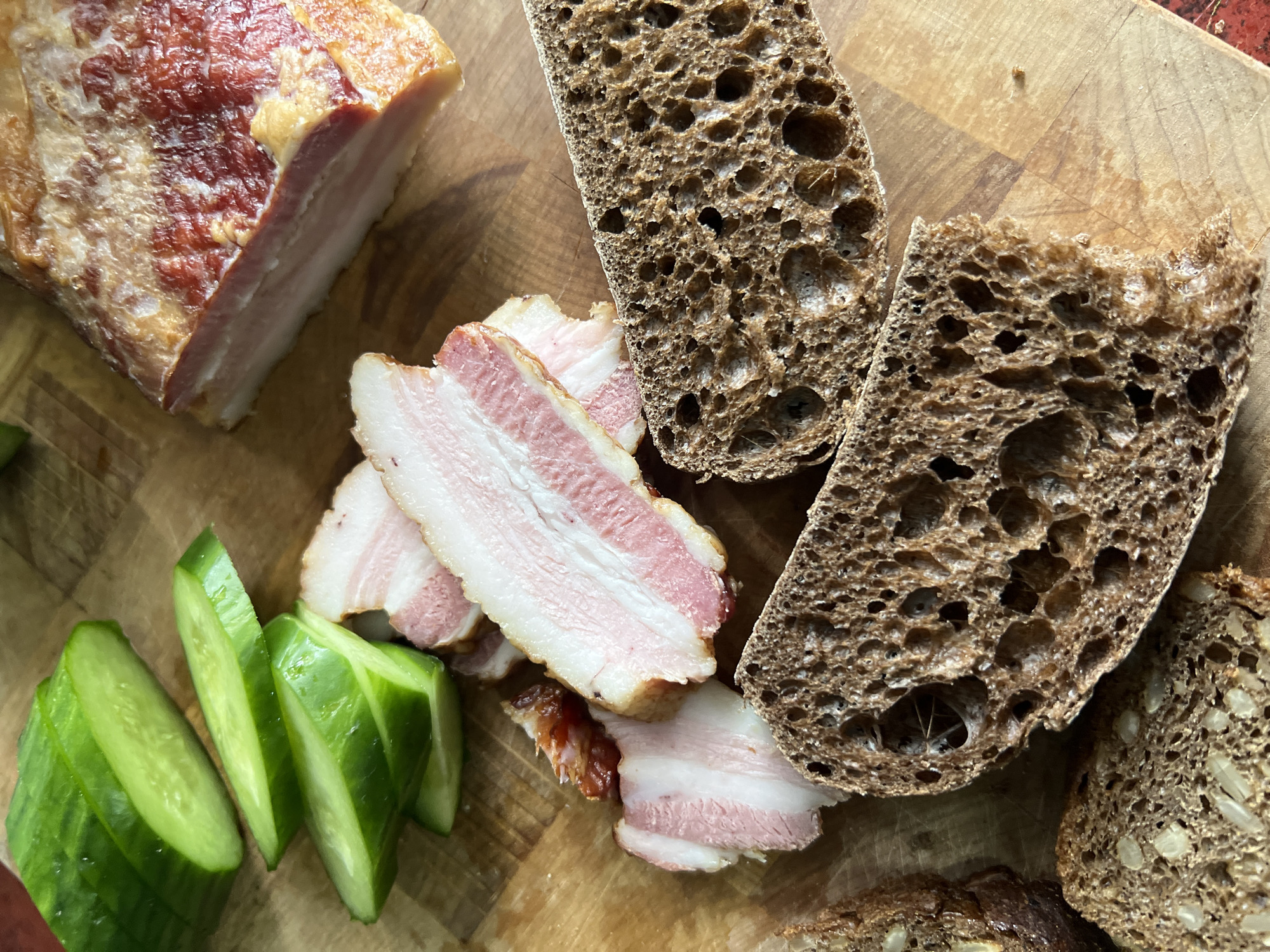 On a wooden cutting board, there is a block of bacon, slices of bacon, sliced cucumbers, and slices of dark rye bread. The image may have been taken in Chaikovsky, Russia.