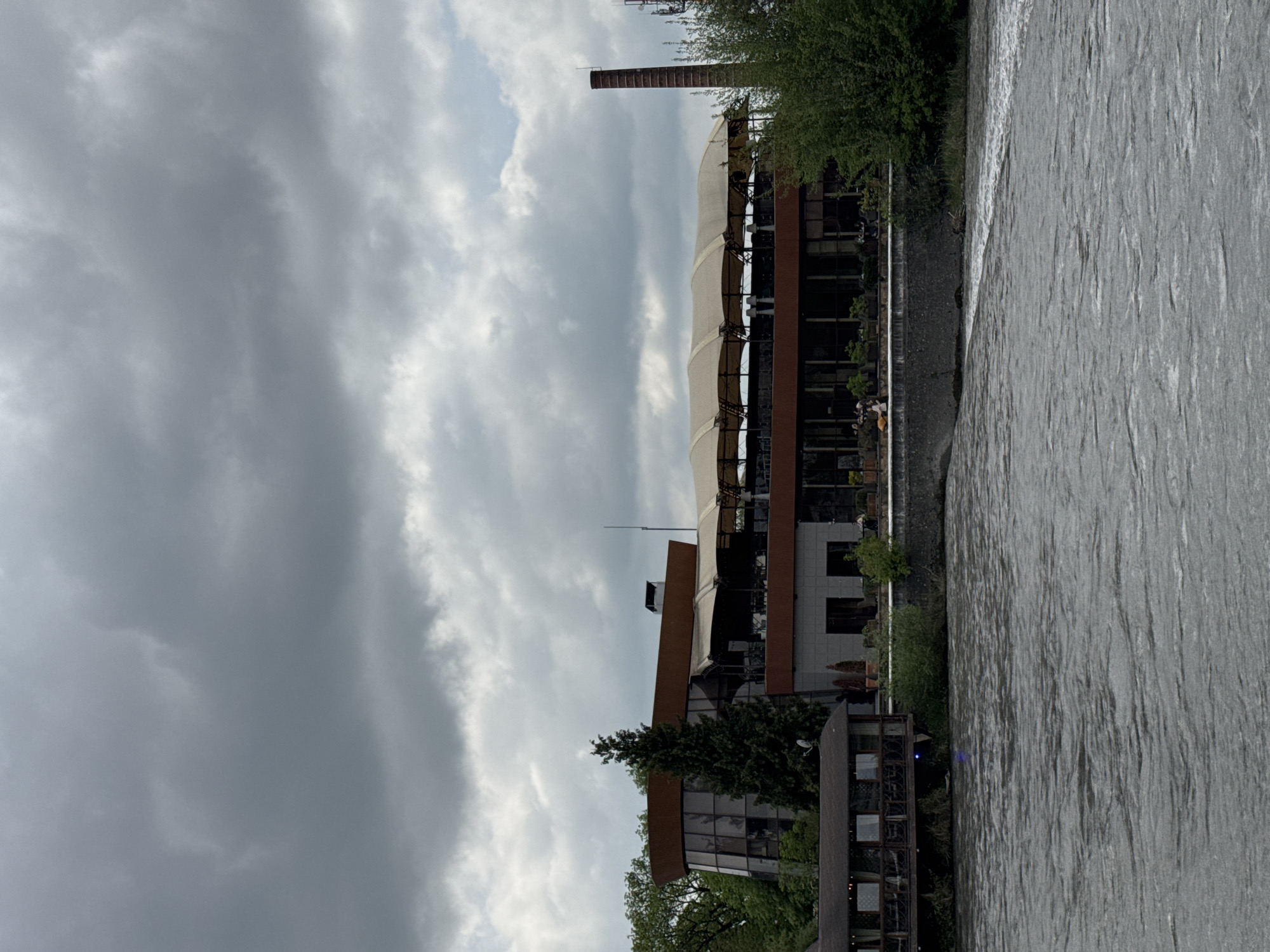 The image depicts a multi-story building situated beside a river. The building has a mix of modern architectural styles, including glass walls, metal framework, and a tent-like roof structure on one section. A tall, brick chimney-like structure stands nearby. The river appears wide and flowing, and its surface shows rippled textures. The sky is overcast, with dark, heavy clouds dominating the scene. Vegetation is visible around the building and along the riverbank, including trees and shrubs. The details provided do not confirm the location in the city of Zamankul, Russia. More forensic information is required.
