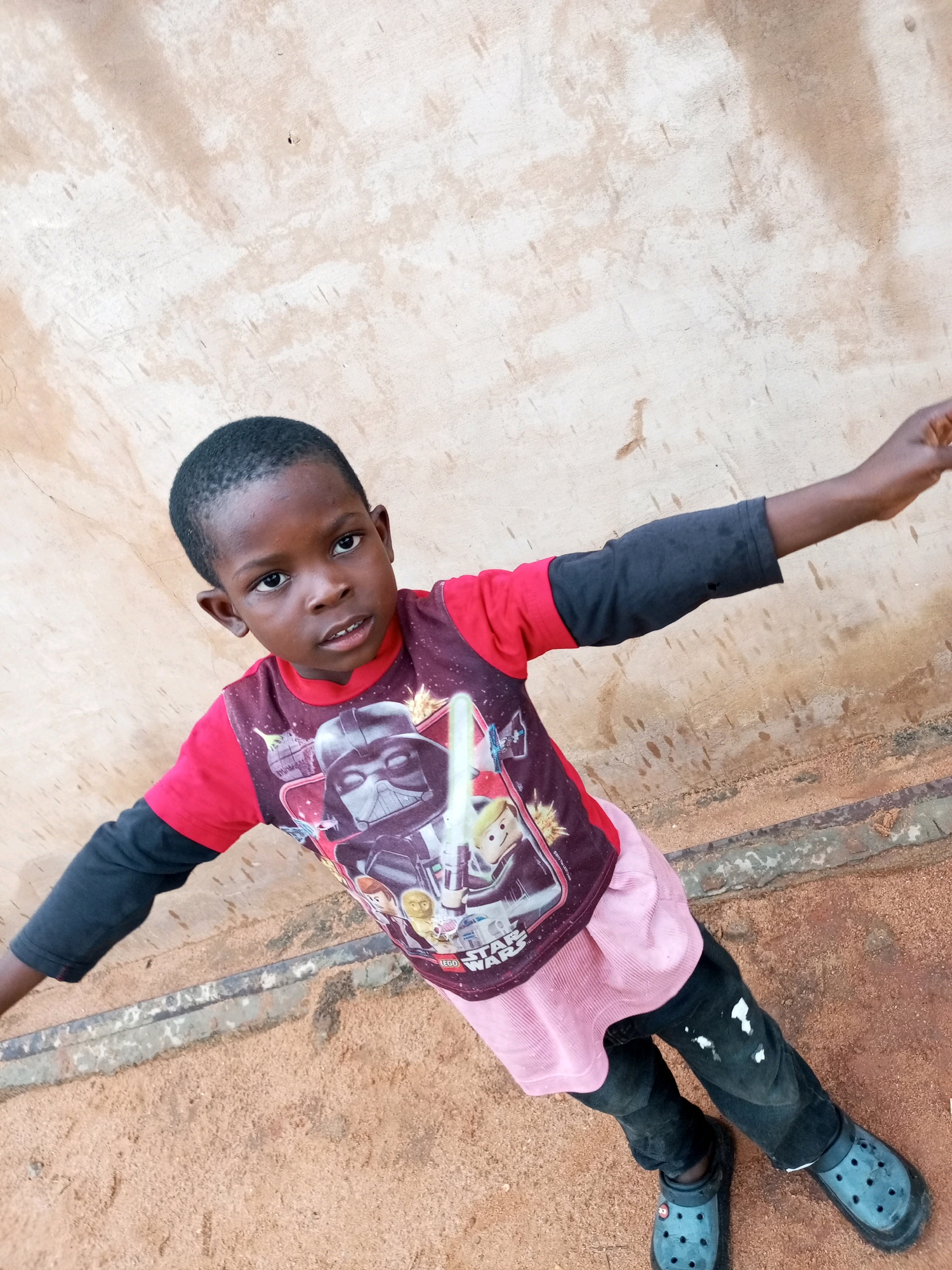 A young boy with dark skin and short black hair stands outdoors against a textured, light brown wall. He wears a red t-shirt with black long sleeves, and a graphic t-shirt over the red one featuring characters from Star Wars, including Darth Vader and Yoda. He has on black pants with white paint splatters and blue Crocs. His arms are outstretched, and he is looking directly at the camera. The ground beneath him is sandy with some debris and a concrete border. The scene appears to be in an outdoor area, possibly a yard or alleyway, in Tempure, Nigeria.