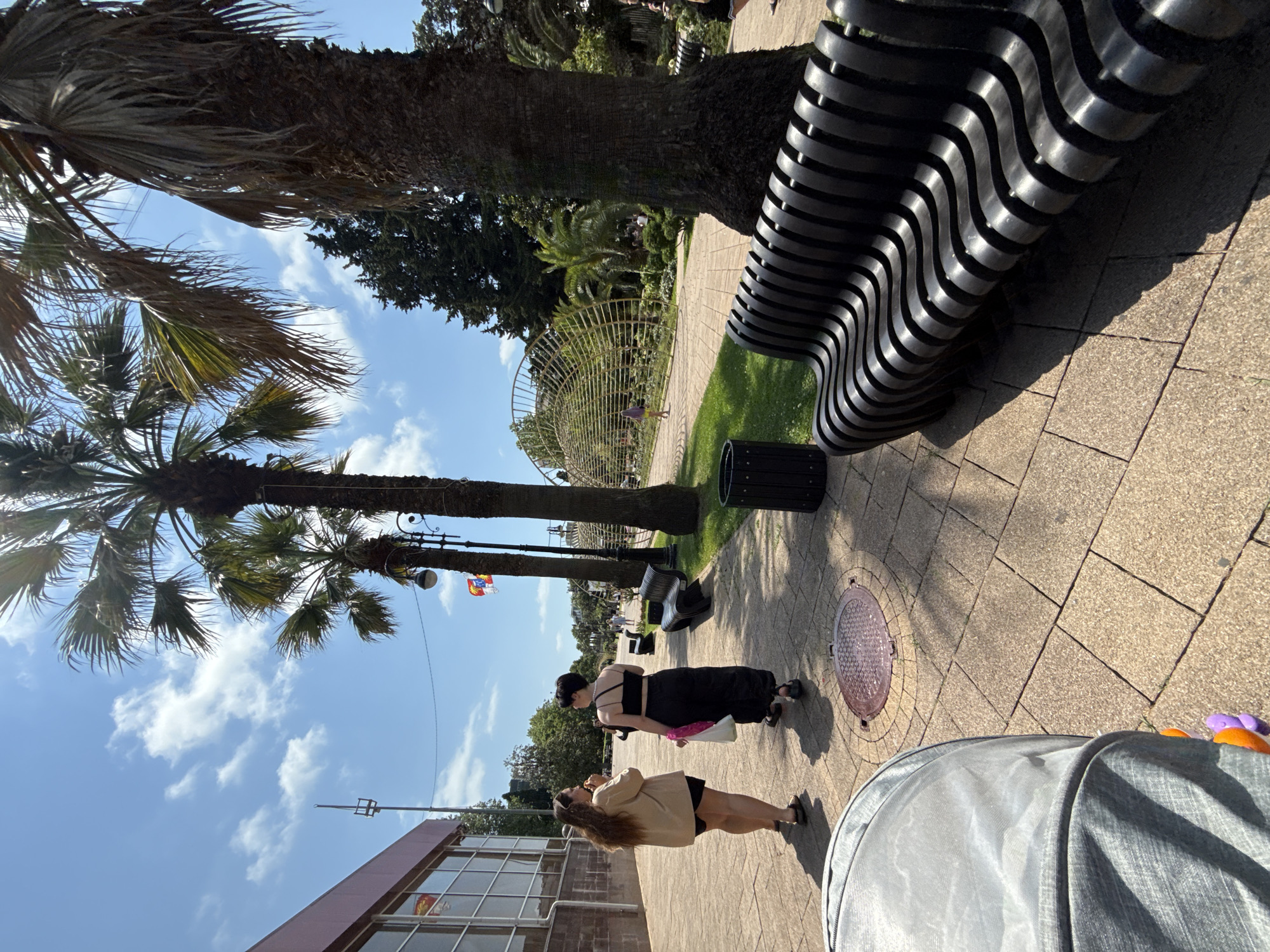 This image depicts an outdoor scene in Sochi, Russia, on a bright, sunny day with scattered clouds. The perspective is angled, giving a sense of looking up and sideways. In the foreground, a person is pushing a stroller, partially visible and covered in a grey fabric.
Further into the scene, two women are walking on a paved pathway. One woman, wearing a black spaghetti-strap top and a long black skirt, is carrying a pink shopping bag. The other woman is wearing a tan jacket over a black outfit and appears to be talking on her phone. Their postures suggest a casual stroll.
The environment features lush greenery with several palm trees, characteristic of a seaside resort. A striking, modern black bench with a wavy design is a prominent object on the right side of the pathway, casting a sharp shadow. Behind the bench, a black trash bin is visible. A circular metal grate is embedded in the pavement.
In the background, a large, open structure with a curved metal framework is partially obscured by trees. A flagpole with a flag, possibly the Russian flag, is also visible in the distance. A building with large windows and a reddish-brown facade is seen on the lower left, indicating an urban or commercial area adjacent to the park-like setting. The lighting suggests it is daytime, likely late morning or early afternoon, given the strong sunlight and distinct shadows. The overall mood appears relaxed and pleasant.