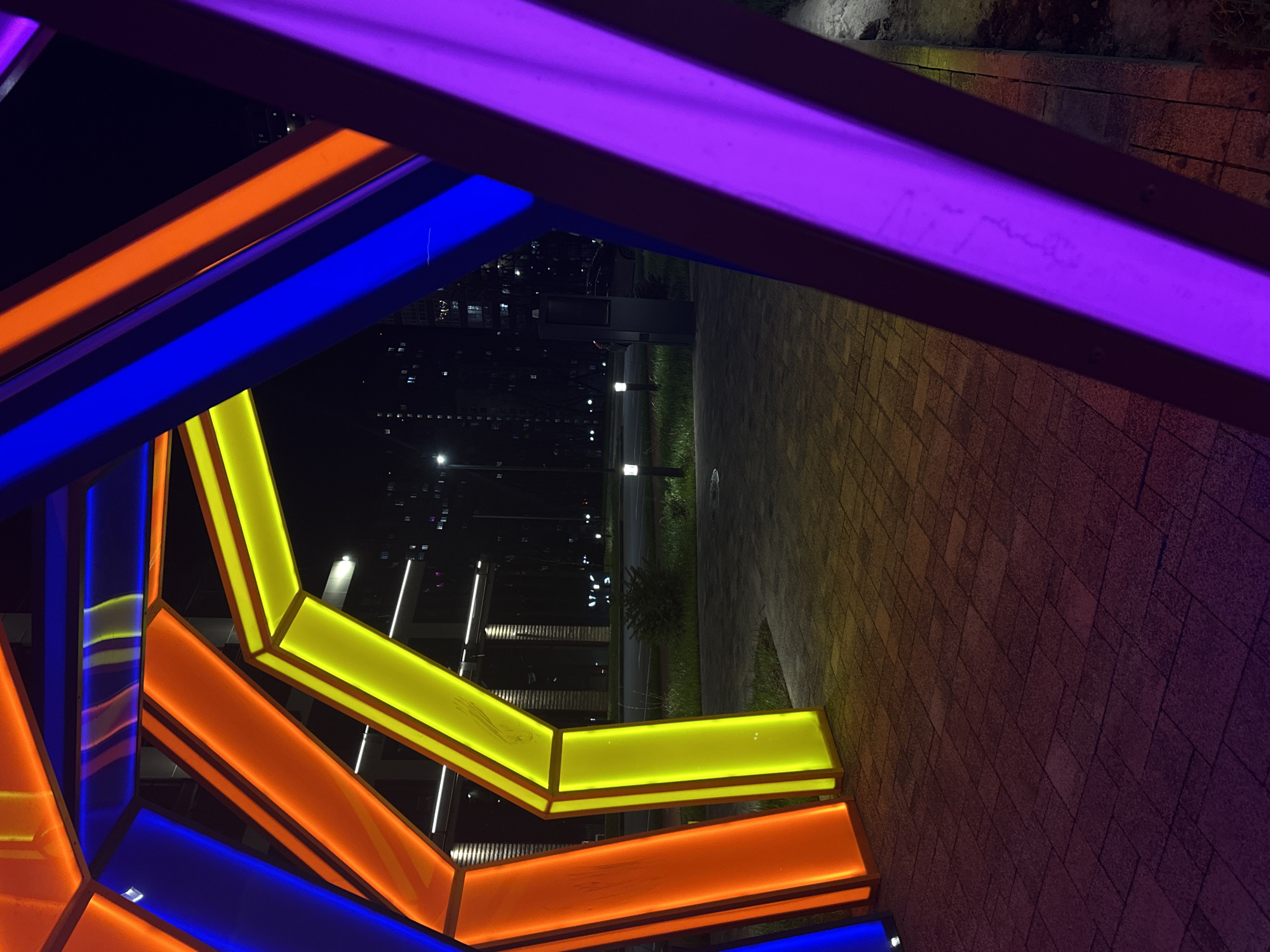 The image shows a nighttime scene in Rostov-on-Don, Russia, featuring a modern art installation made of illuminated rectangular prisms in blue, orange, yellow, and purple colors. The installation is located outdoors, adjacent to a paved walkway or plaza. In the background, streetlights line a road, and several multi-story buildings with illuminated windows are visible. The photo is taken from a low angle, looking up through the art installation toward the cityscape.