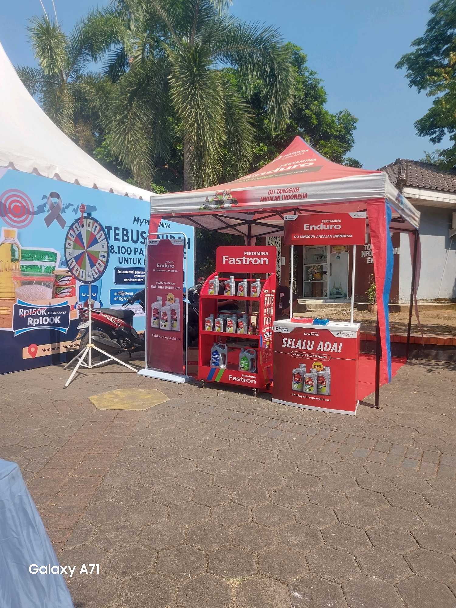 This is an outdoor scene in Makassar, Indonesia, under a clear blue sky with palm trees and other foliage in the background. In the foreground, there is a promotional booth set up, likely for a fuel or oil company. The booth features red and white tents. To the left of the main booth, there is a large blue banner with promotional graphics including a spinning wheel, images of food items like rice and cooking oil, and text in Indonesian. A red motorcycle is parked near the banner. The central part of the scene is dominated by a red promotional kiosk or stall with various oil products displayed. On the right side of the kiosk, there is a tall red display rack filled with different types and sizes of oil bottles, branded as "Fastron". The kiosk itself has signage with "ENDURO" and "SELU ADA!" (meaning "Always Available!"). The ground is paved with interlocking hexagonal stones. The overall impression is of a promotional event or roadside stall.