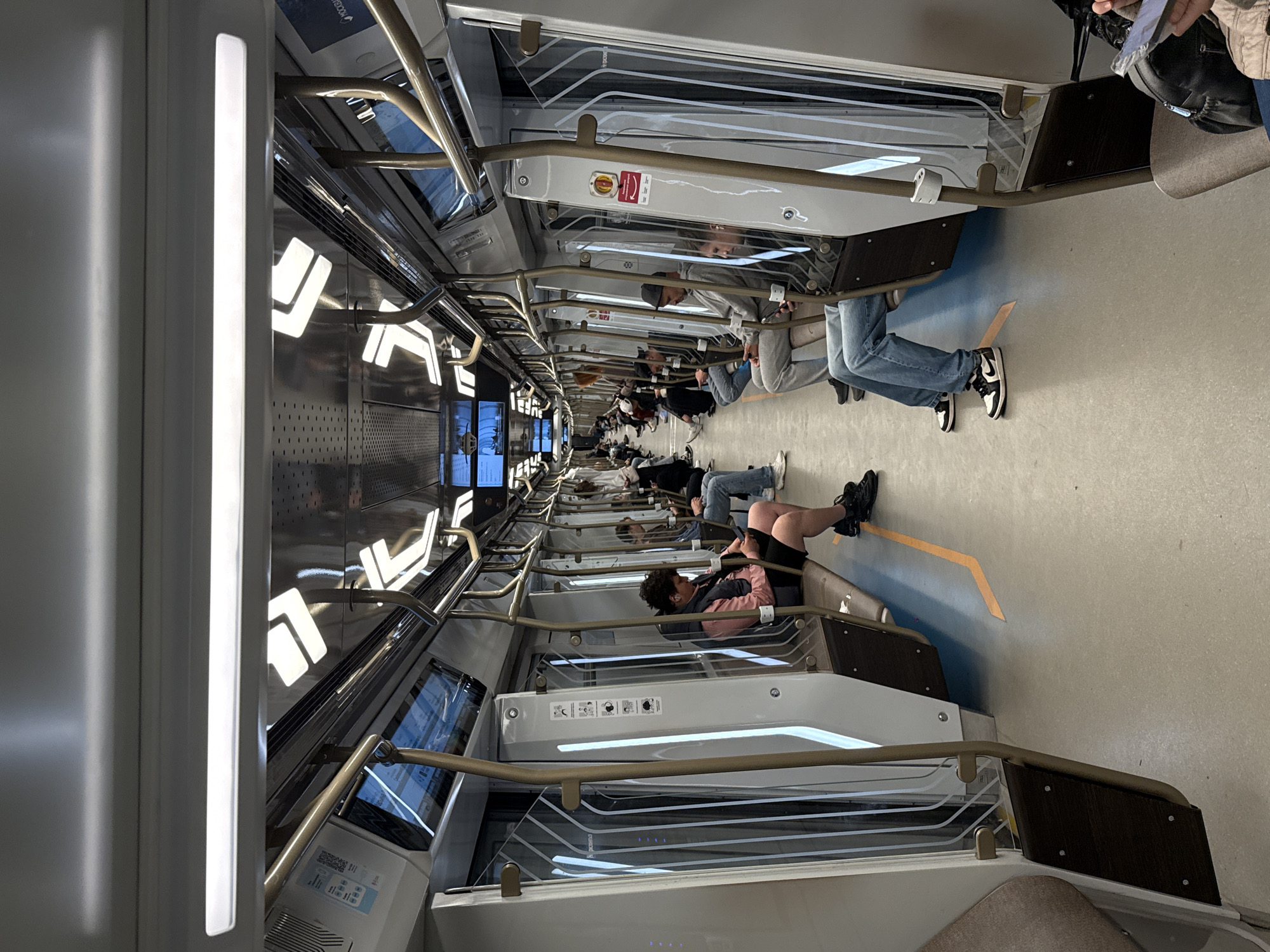 The image captures the interior of a modern subway train car, seen from a perspective looking down the length of the carriage. The car is sleek and minimalist, featuring light gray and white surfaces, with integrated strip lighting running along the ceiling and walls.
Numerous passengers are visible, primarily young adults, occupying the specialized seating arrangements. Unlike traditional subway seats, these appear to be padded ledges or benches along the walls, allowing people to face inwards into the aisle. Many individuals are taking advantage of this design to recline or lie down comfortably, with some stretched out, resting their heads against the wall and their feet extended onto a blue and yellow marked section of the floor. Other passengers are sitting upright, looking at their phones or resting. The general atmosphere appears calm and relaxed, suggesting a comfortable journey, possibly during off-peak hours or a longer route.
Notable details include robust, golden-colored grab bars running horizontally above the seating areas, and vertically from the ceiling. Digital display screens are embedded along the ceiling/wall, with one partially showing text that includes "VOZDUX." Small informational icons and text are visible on panels next to some seating sections, likely providing instructions or warnings, including a red emergency sign. Reflections of illuminated angular patterns are visible on the metallic wall panels above the windows. The flooring is a light, uniform color. The modern design is characteristic of newer trains, such as those found in the Moscow Metro.