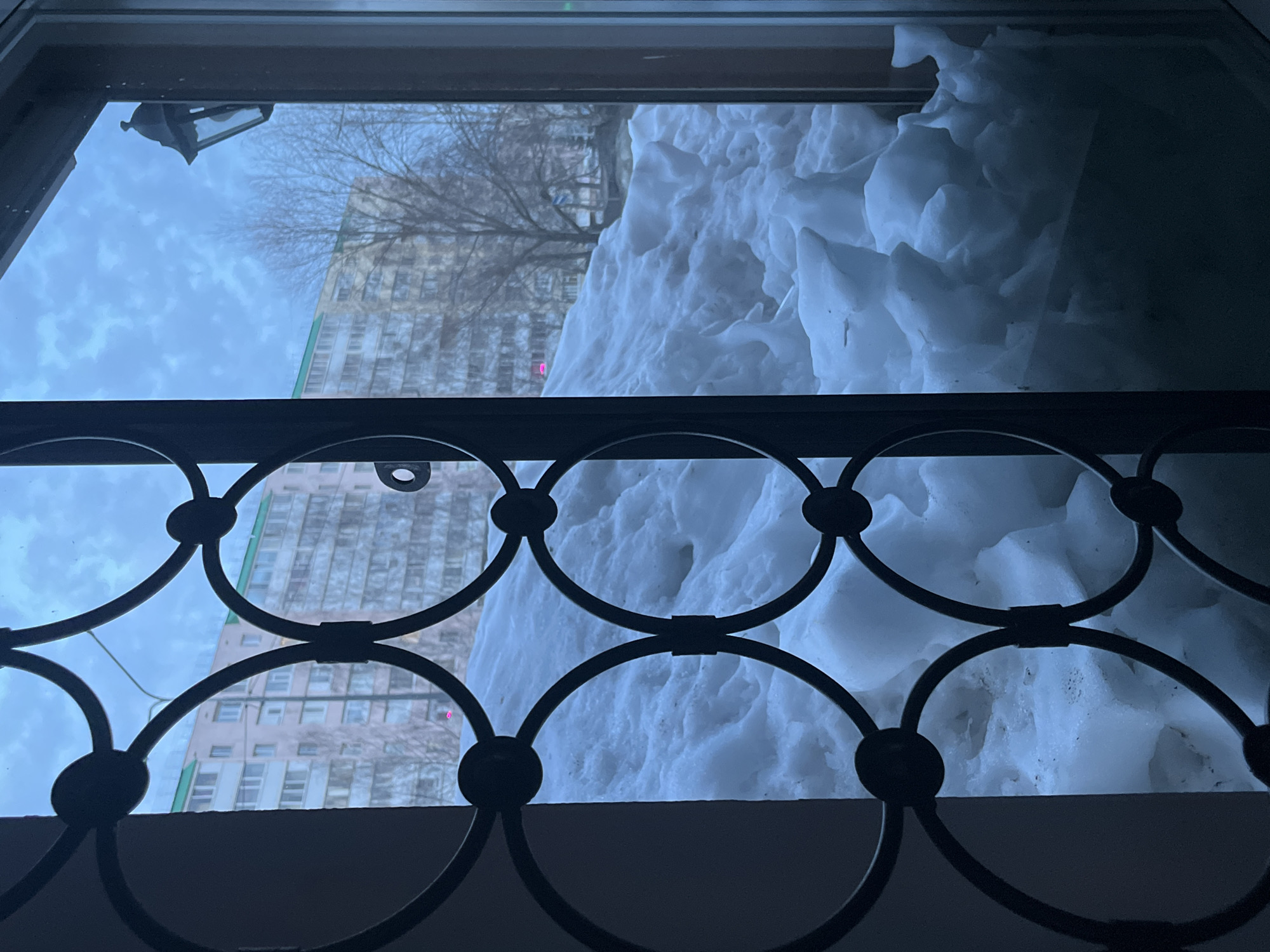 The image depicts an exterior urban scene in Neftekamsk, Russia, viewed through a dark, decorative metal-grated window or door. The grating features a pattern of interconnected circular and curved elements. Immediately outside, a large, irregularly shaped mound of snow occupies the lower and central portions of the view, extending upwards to approximately the mid-point of the frame. The snow exhibits varied textures, indicating accumulation and possibly partial melting/refreezing. Beyond the snow, a sparsely treed area with bare deciduous trees is visible. In the background, multiple multi-story residential buildings with light-colored facades, some featuring green roof details, stand under an overcast, light blue-grey sky. A black, antique-style lantern is affixed to an unseen structure in the upper right quadrant of the frame. No individuals, explicit actions, or interactions are present in the scene.
