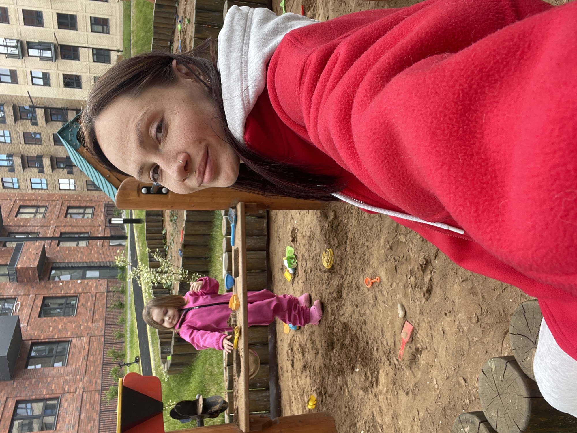 The image presents a medium shot of a woman wearing a red hooded jacket with a white hood. She is looking directly at the camera and is positioned in the foreground, partially obscuring the background. In the background, there is a child in a pink outfit playing in a sandbox. The sandbox is surrounded by wooden structures, and there are toys scattered in the sand. Further in the background is a building with multiple windows. The location is Pervomaysky, Russia.