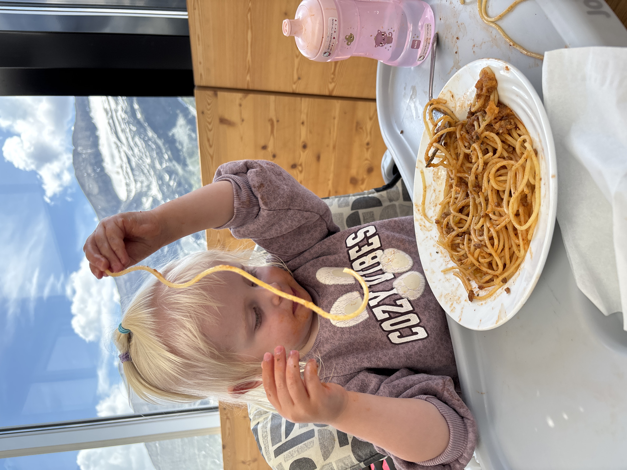 A young child with blonde hair pulled into pigtails with blue and purple ties is sitting in a high chair and eating spaghetti. The child is wearing a purple sweatshirt with "Cozy Vibes" printed on it in white letters. A plate of spaghetti is in front of the child, and they are holding a strand of spaghetti up to their mouth. To the right of the plate is a pink sippy cup. In the background, there is a window with a view of mountains and a cloudy sky. The scene appears to be in a restaurant in Heiligenblut am Großglockner, Austria, as indicated by the background scenery which is characteristic of the region.