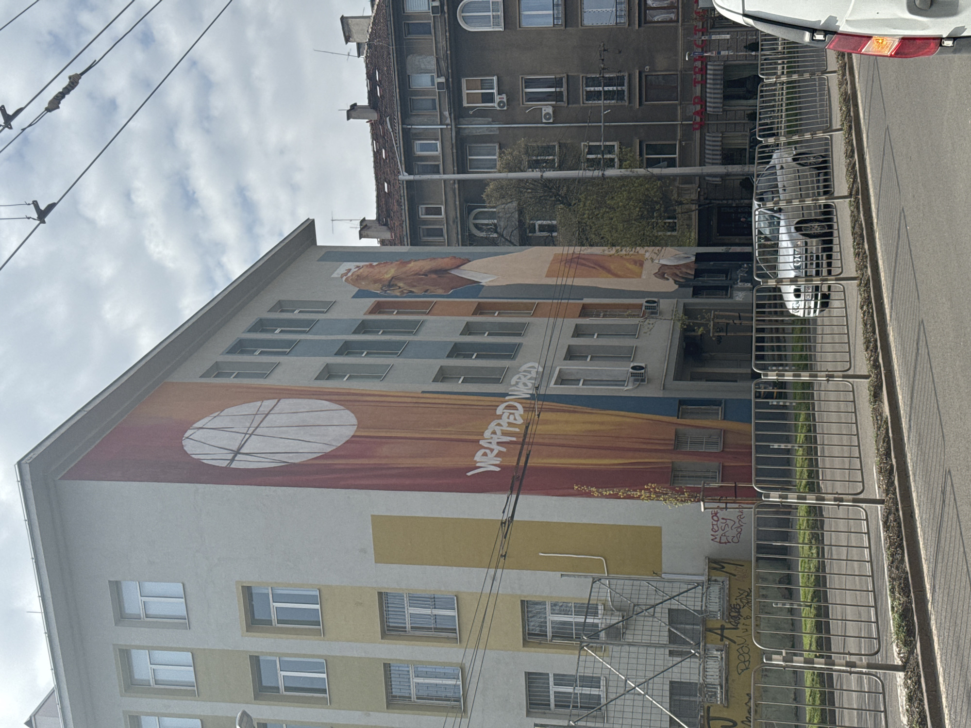 The image shows a building in Sofia, Bulgaria. The building features a large mural that covers several stories. The mural includes a circle with lines across it, text reading "WRAPPED WORLD," and an image of a hand. The building is situated on a street with a fence running along the sidewalk. Across the street is another building with multiple windows. A white vehicle is partially visible in the upper right corner of the image. The sky is overcast.