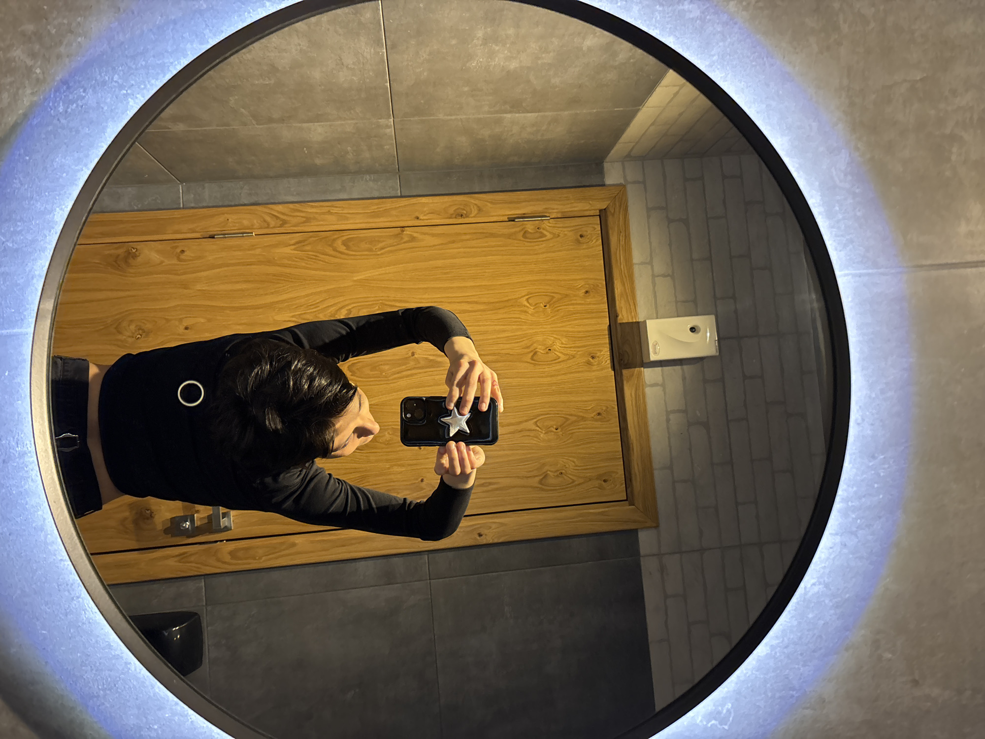 A person is taking a selfie in an illuminated round mirror in a bathroom. The person is wearing a black long-sleeved shirt and holding a black smartphone with a silver star attached to the back. The bathroom features grey tile flooring, a wooden door, and white brick-patterned tiles on the wall. A white soap dispenser is mounted on the wall. The location is Sofia, Bulgaria.