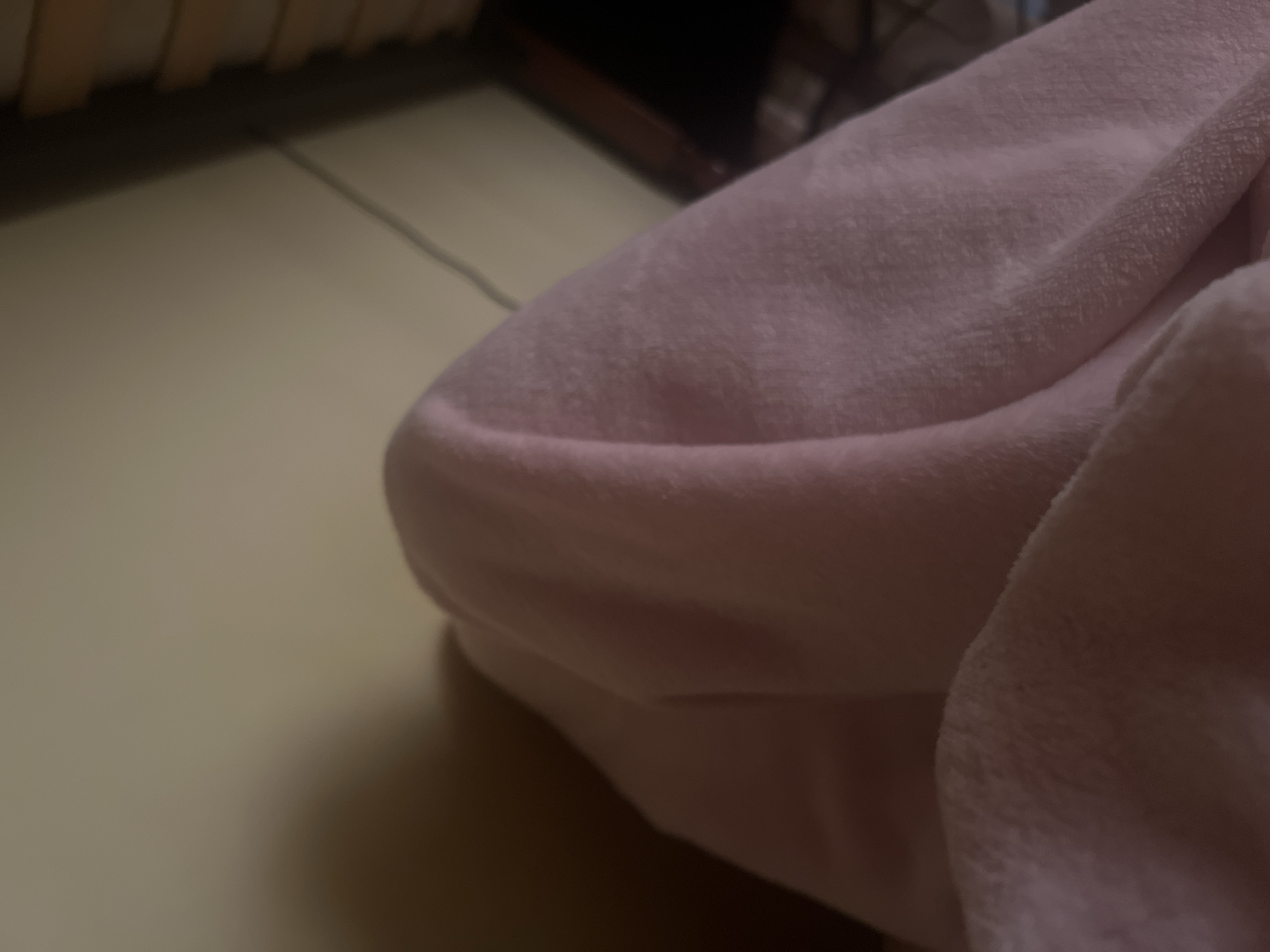 A close-up shot focuses on a pink fuzzy blanket draped over an unseen object, creating a soft, rounded shape. The blanket's texture is visible, with a slightly matted appearance in places. Behind the blanket, a wall with a subtle gradient of light and shadow can be seen. To the right, a dark, out-of-focus background suggests furniture or a bookshelf. A thin black cord runs diagonally across the upper right portion of the frame. The lighting is dim, casting a soft glow on the blanket and creating a shadow on the wall. The scene appears to be indoors, possibly in a bedroom or living area, with no specific city or location details discernible from the image.