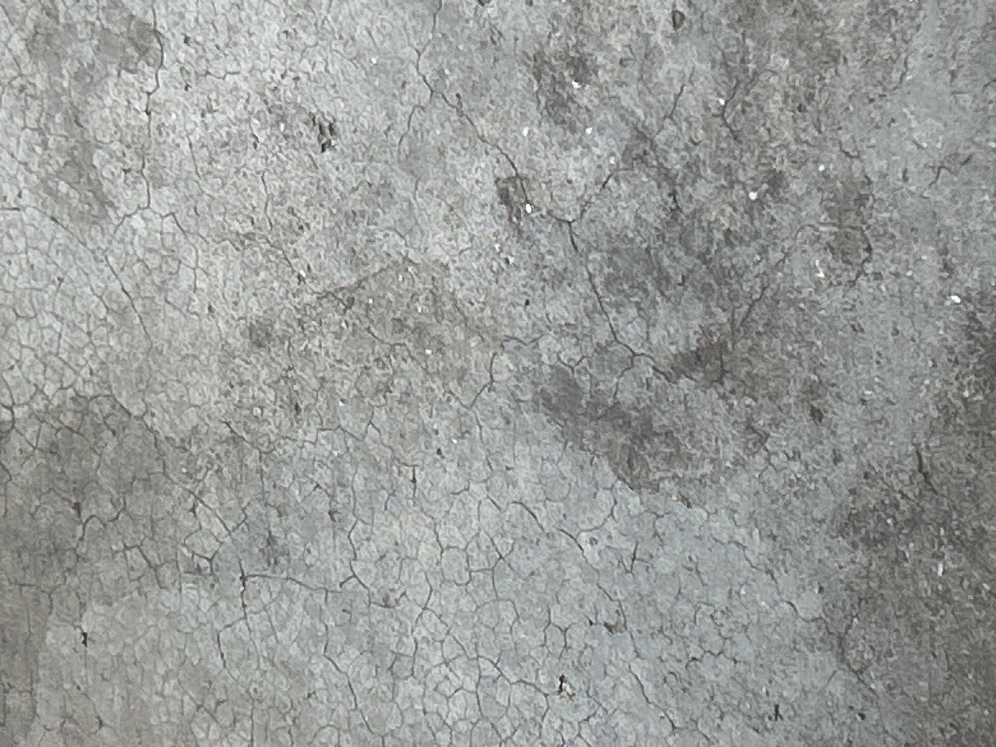 The image displays a close-up, overhead view of a cracked concrete surface. The concrete exhibits a weathered, grey texture with numerous fine, dark lines forming a network of cracks across the entire frame. The cracks vary in size and density, with some areas showing more fragmentation than others. There are also some darker patches and lighter flecks scattered throughout the surface, suggesting variations in the concrete mixture or surface debris. The lighting appears to be diffuse, casting no distinct shadows. The image does not contain any people, animals, or discernable objects other than the concrete itself.
This image depicts a detail of an outdoor surface, likely a pavement, sidewalk, or floor, which could be found in various locations within the city of Santa Ana, Venezuela, or any other urban or rural area with concrete infrastructure. Without additional context or identifying features, it is impossible to pinpoint a specific location within Santa Ana.