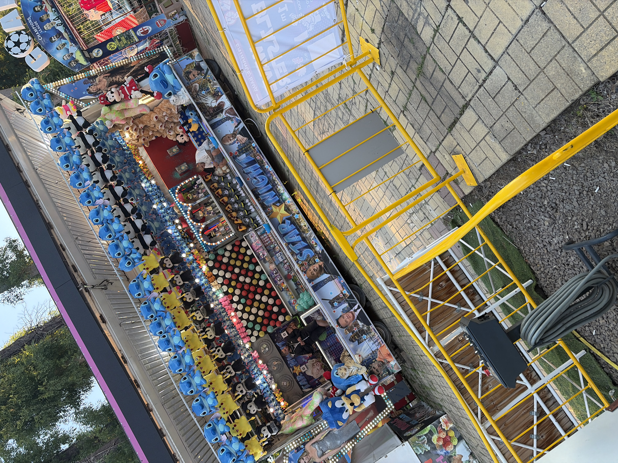 The image captures a close-up, tilted perspective of an amusement park or carnival game stall. The stall is adorned with numerous plush toys, primarily blue Stitch characters from Disney's Lilo & Stitch, yellow Pikachu, and various other cartoon figures, hanging in rows. Below the toys, the stall displays images of celebrities and athletes, including what appear to be actors and basketball players, along with a banner that reads "STAR NIGHT."
The stall itself is decorated with a bright, colorful aesthetic, featuring a red and yellow striped canopy. In front of the stall, a bright yellow metal barricade is positioned, with a gray electrical control box and a coiled black cable situated on the ground to the left of the barricade. The ground is a mixture of paved brickwork and gravel.
In the background, lush green trees and a clear blue sky are visible, suggesting an outdoor setting. The overall scene depicts a vibrant, playful atmosphere typical of a fairground attraction.