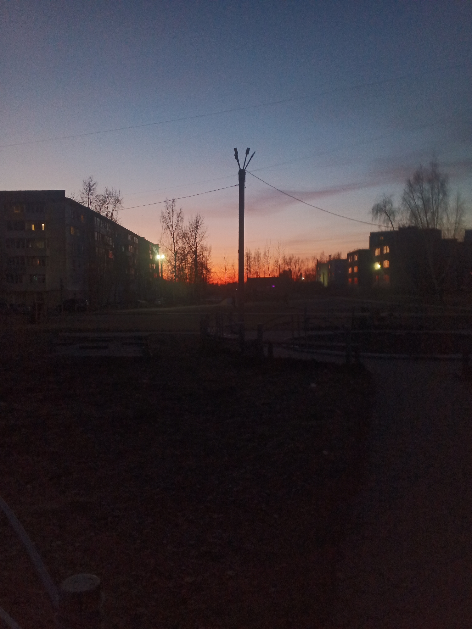 This image depicts an urban or suburban landscape in Chaikovsky, Russia, captured during twilight, showcasing a striking sky at either sunrise or sunset. The sky transitions dramatically from a deep dark blue at the top to vivid oranges and reds along the horizon line.
In the foreground, a dark, unpaved ground area suggests a yard or undeveloped space. A tall, dark utility pole with a multi-pronged light fixture stands prominently in the midground, with power lines stretching across the upper portion of the frame.
Multi-story residential buildings frame the scene. On the left, a large apartment block with an architectural style consistent with Soviet-era construction shows many windows, several of which are illuminated with warm yellow light. A bright street lamp also casts light near this building. On the right side, other buildings are mostly silhouetted against the colorful sky, also revealing a scattering of lit windows. Numerous bare trees, typical of late autumn or winter, are visible between and behind the structures, their branches silhouetted against the horizon.
No people or overt activities are visible, contributing to a quiet and serene atmosphere. The clear sky indicates calm weather. The overall low light levels emphasize the artificial illumination from the buildings and the natural, vibrant hues of the twilight sky. There is no visible text in the image.