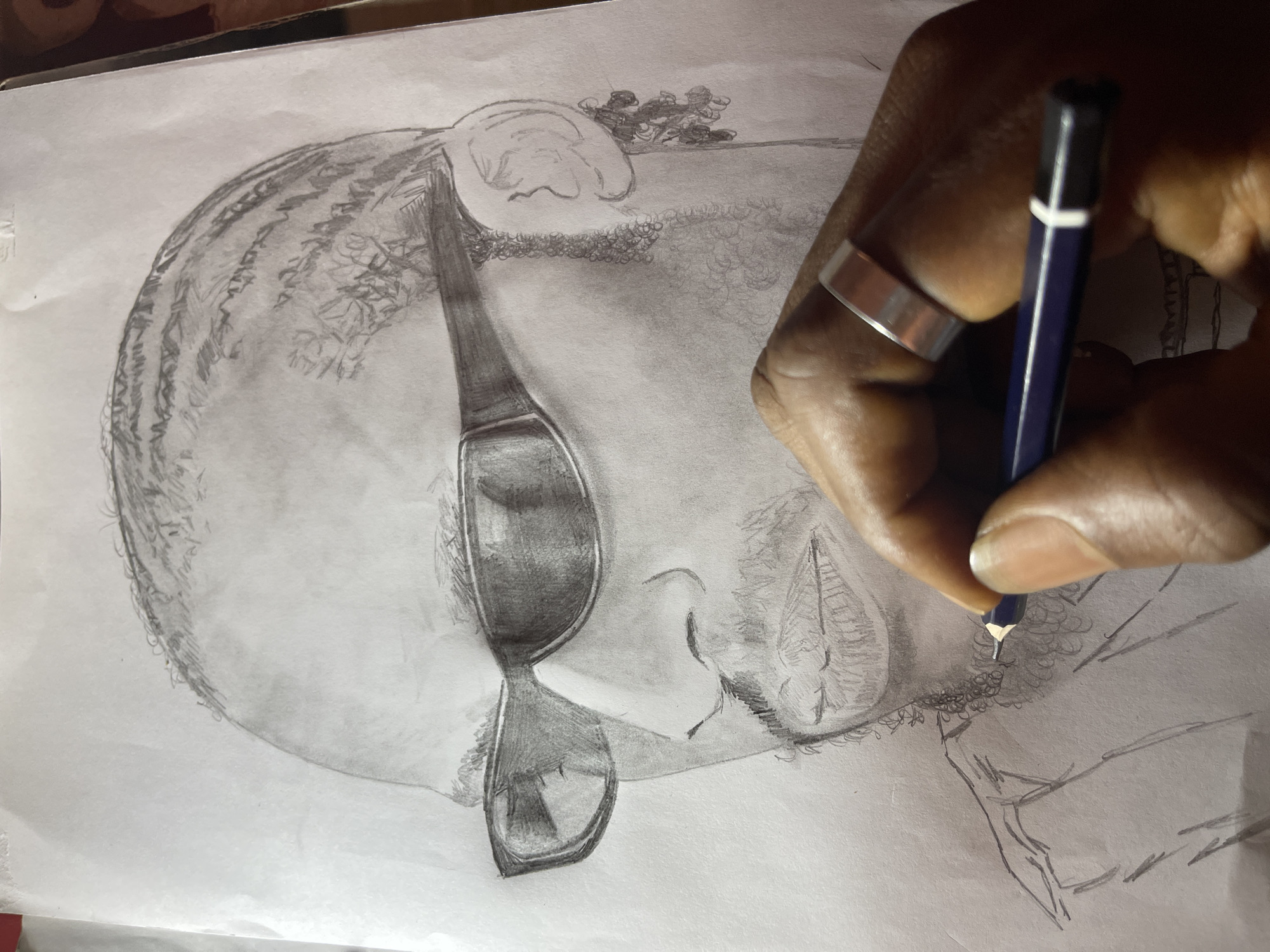 A close-up photograph depicts a pencil drawing of a man's portrait in progress on a sheet of white paper. The drawing, rendered in various shades of graphite, showcases a male figure in a three-quarter profile facing left, wearing dark, rectangular sunglasses that obscure his eyes. He has discernible facial features including a nose, closed lips, and a short, textured beard and mustache. His hair is styled in distinct cornrows or dreadlocks that sweep backward from his forehead. The upper torso of the subject is partially outlined, indicating an unfinished section of the drawing.
In the lower right portion of the frame, a dark-skinned hand, presumably the artist's, is actively holding a dark blue pencil with a visible sharpened tip. The pencil is positioned to add detail to the man's beard area. A metallic, reflective band-style ring is worn on the ring finger of the hand. The paper exhibits minor creasing near the upper edge. The scene, depicting this artistic activity, is situated in the city of Jos, Nigeria.