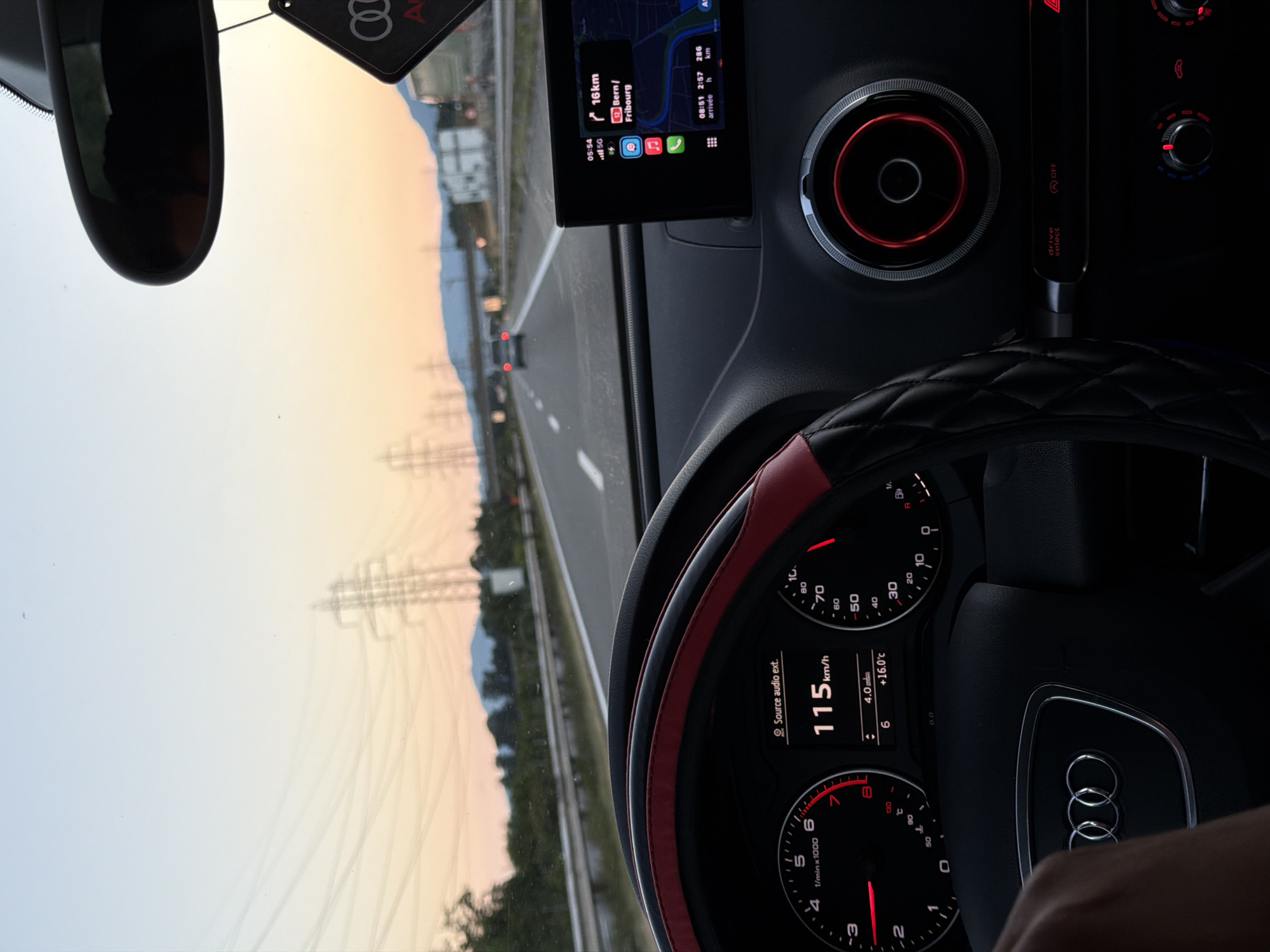 The image shows the interior of a car, specifically the dashboard and steering wheel, as seen from the driver's perspective. The car is on a highway, with a sunset visible through the windshield. The dashboard displays the car's speed at 115 km/h and the outside temperature at +16.0°C. A navigation screen shows a route to Bern/Fribourg, indicating a distance of 16 km. The car appears to be an Audi, based on the emblem on the steering wheel and the design of the air vents. Electrical pylons and trees line the side of the highway. The scene is set during dusk, with a warm, orange hue in the sky. The location is identified as Yvorne, Switzerland.