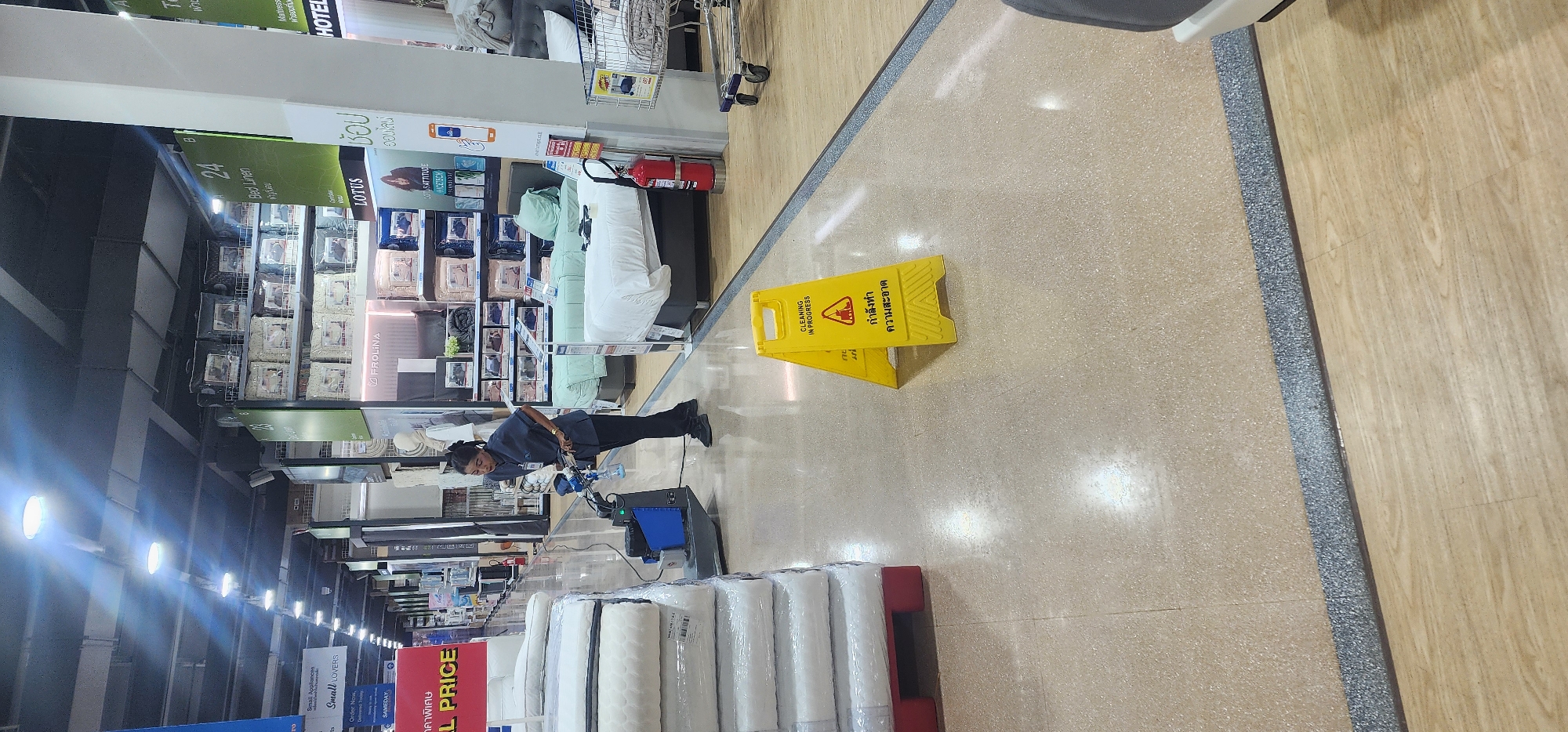 In a retail store in Baan Chaweng Noi, Thailand, a female employee is operating a floor cleaning machine. The employee, wearing a dark blue uniform, is positioned behind the machine, which is emitting water and appears to be scrubbing the floor. A yellow "Wet Floor" sign with Thai text indicating "Cleaning in Progress" is prominently displayed on the floor in the foreground, warning of the wet conditions. The store environment features aisles with merchandise, including stacked mattresses wrapped in plastic on the left side of the image and displayed bedding on shelves in the background. The floor is a light-colored, speckled surface, with sections of light wood-grain flooring also visible. Lighting fixtures are visible on the ceiling.