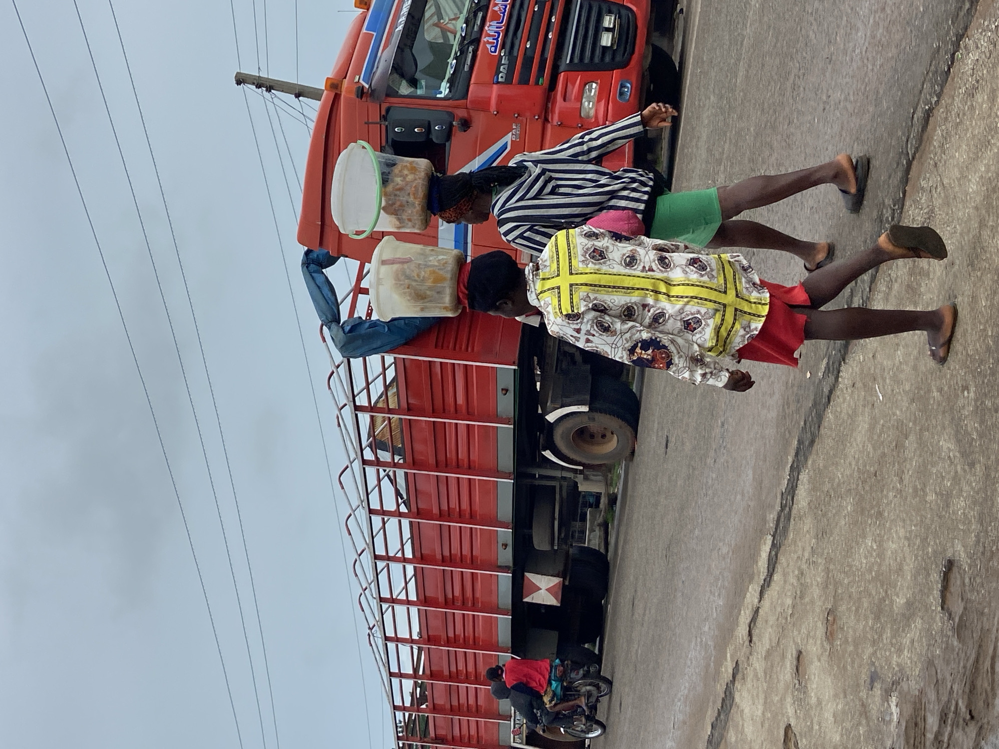 The image depicts a street scene in Mararaba, Nigeria, under an overcast sky. A large red flatbed truck, identified by Arabic script reading "ما شاء الله" on its cab, dominates the background. The truck features orange warning lights on its cab and a blue tarp secured to its flatbed, which has tall red railings and a white and red 'X' hazard marker on its rear lower section.
In the foreground, two women walk away from the viewer on the right side of the road. Both women are dark-skinned and wear sandals. The woman on the left wears a white long-sleeved shirt with a yellow cross-like pattern on the back and red shorts; she carries a clear plastic bucket filled with light-colored food items on her head, secured by red fabric. The woman on the right wears a black and white striped long-sleeved top and green shorts; she also carries a clear plastic bucket of light-colored food on her head, secured by dark fabric, and has a pink bag slung over her left shoulder.
To the left, partially obscured by the truck, a dark-colored motorcycle transports two individuals. The front rider wears dark clothing, and the passenger wears a red shirt. Overhead, multiple thin utility lines stretch across the grey, featureless sky. The road surface is a worn, cracked grey pavement.
