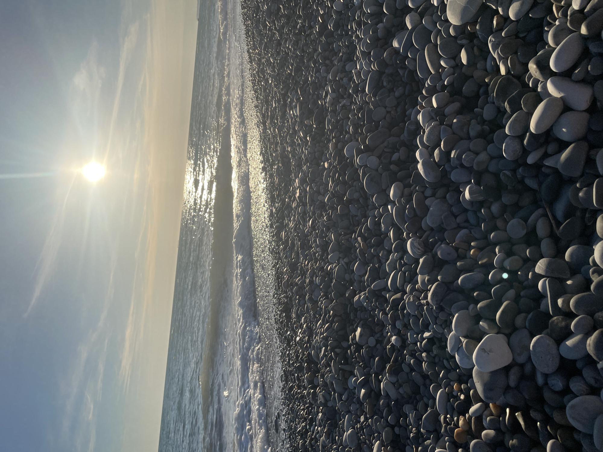 This media file depicts a serene beach scene at sunrise. The image is oriented vertically, with the sun low in the sky, casting a warm, golden glow and creating a shimmering reflection on the water's surface. Wispy clouds are visible in the bright sky. To the right of the frame, a vast expanse of smooth, grey pebbles covers the beach, with some lighter-colored stones interspersed among them. The pebbles are densely packed and cast long shadows, indicating the low angle of the sun. Small waves gently lap at the shore, their edges marked by white foam. The overall mood is peaceful and tranquil, evoking a sense of calm and natural beauty. There is no visible text in the image.