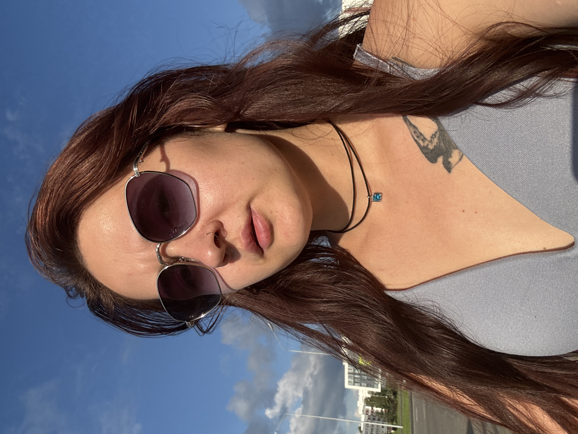 A medium shot captures a woman with long, dark auburn hair from the chest up, standing outdoors against a bright blue sky with scattered clouds. She is wearing oversized, silver-rimmed sunglasses with gradient lenses, a thin black choker necklace with a small, square blue pendant, and a light blue, V-neck top. A bird tattoo is visible on her upper chest. The background shows a flat, paved area, possibly a street or parking lot, with modern buildings and streetlights on the left side. The scene appears to be in the city of Naberezhnye Chelny, Russia, based on the architectural style of the buildings.