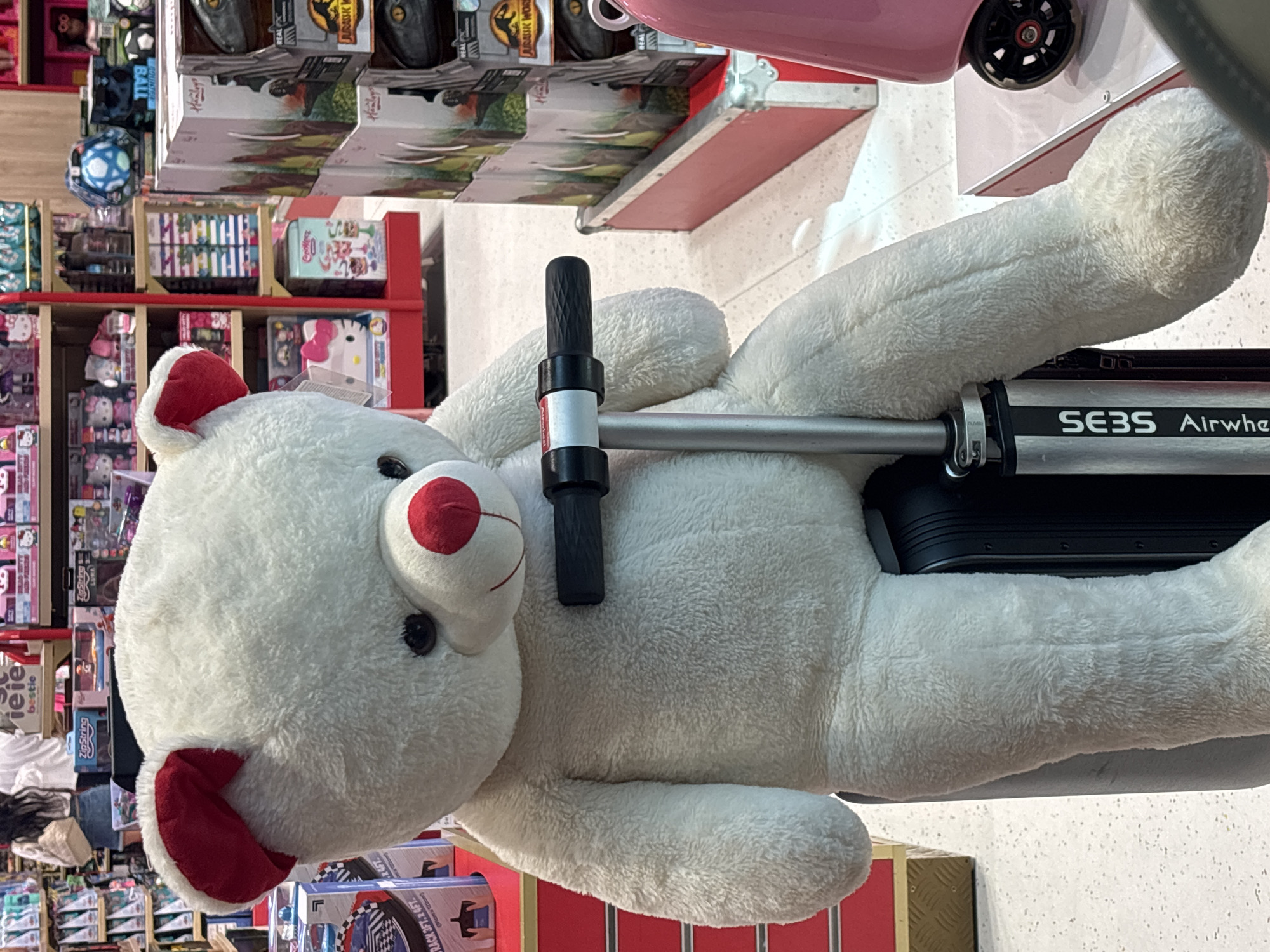 A large, fluffy white teddy bear with red ears and a red nose is sitting on a black suitcase with a silver scooter handlebar resting on its chest. The teddy bear is positioned centrally in the frame, facing forward. Behind the teddy bear, shelves are stocked with various toys and merchandise, including Hello Kitty items and Jurassic World dinosaur toys. To the right of the teddy bear, a pink suitcase with a black wheel is partially visible. The scene appears to be inside a retail store. The image quality suggests it was taken with a mobile phone.