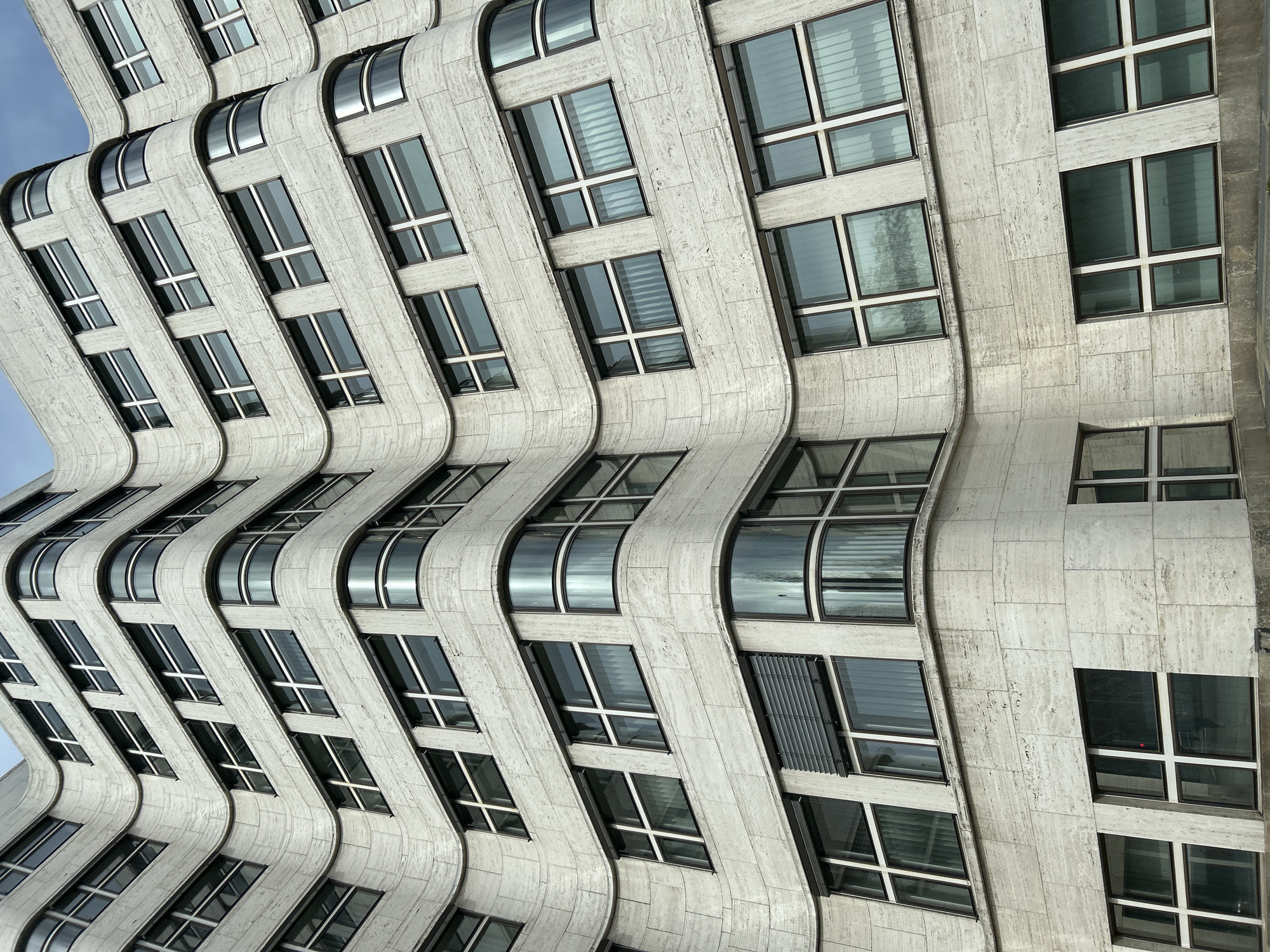 The image shows a section of the undulating facade of the Neuer Zollhof building in Düsseldorf, Germany. The architectural feature is characterized by curved, white-grey stone elements and rectangular windows with a white grid. The windows reflect the sky and surrounding structures. Some windows are covered with blinds. The angle of the shot is upward. A small portion of the blue sky is visible in the upper-left corner.