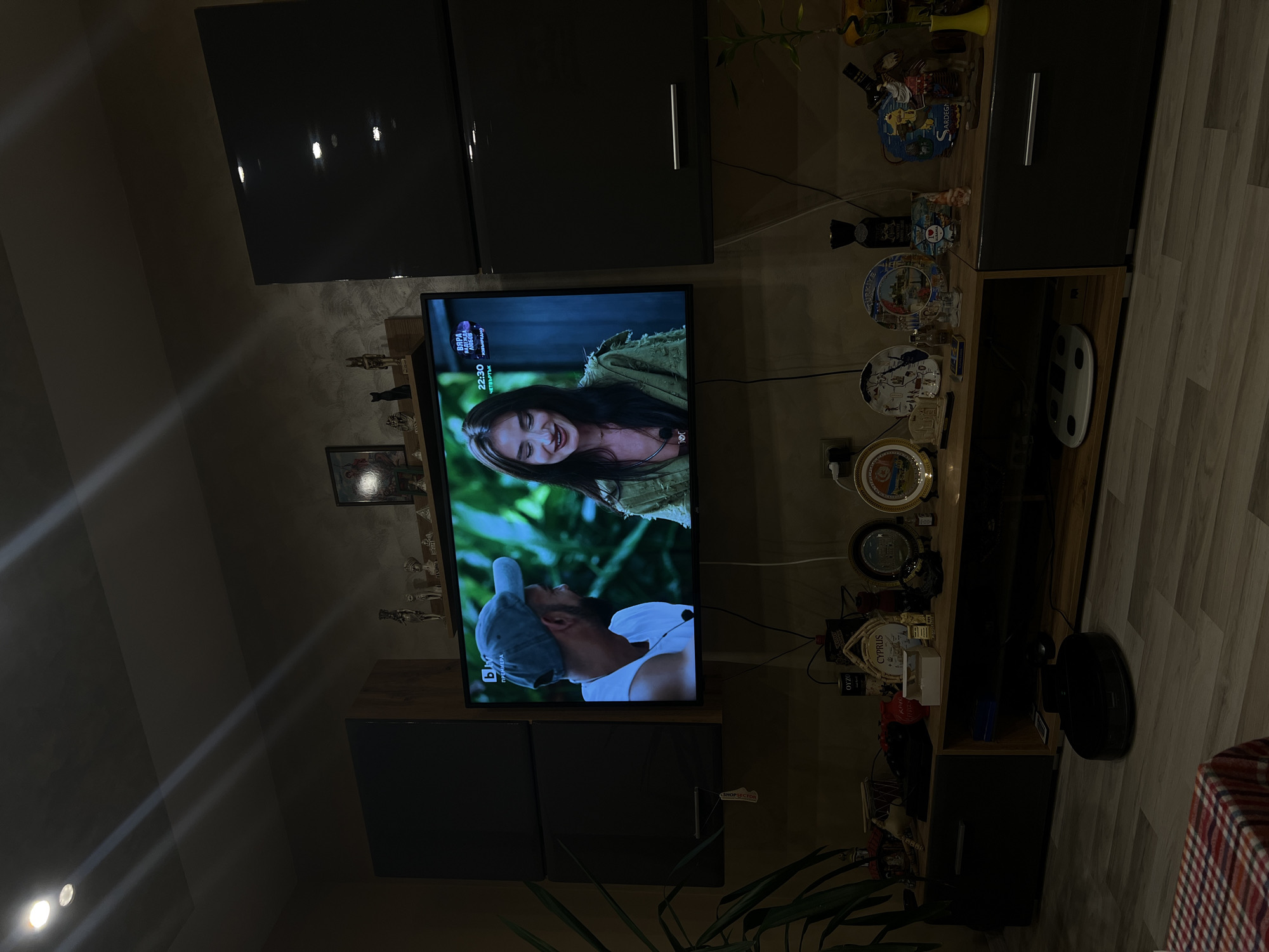 The image shows the interior of a home, likely in Popintsi, Bulgaria. The focal point is a television displaying a scene with a woman and a man. Above the TV are dark-colored cabinets. To the right is a shelving unit displaying various decorative items, and a floor lamp is positioned nearby. A vacuum cleaner is visible on the floor, and a plant is located in the lower portion of the photo. The scene is dimly lit. The flooring appears to be laminate.