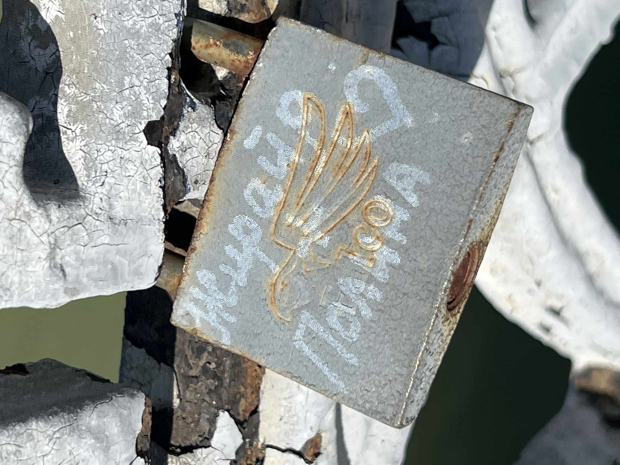 A close-up shot depicts a gray, square-shaped padlock secured to a white, possibly metal, structure. The padlock features a raised trident-like symbol in its center, with white Cyrillic text painted around it. The padlock shows signs of wear and rust, particularly around its edges and lock mechanism. The structure to which it is attached also displays aging, with cracked and peeling paint. The background is blurred, but it appears to be a natural setting with green and white elements. The location is in the city of Krasnodar, Russia.