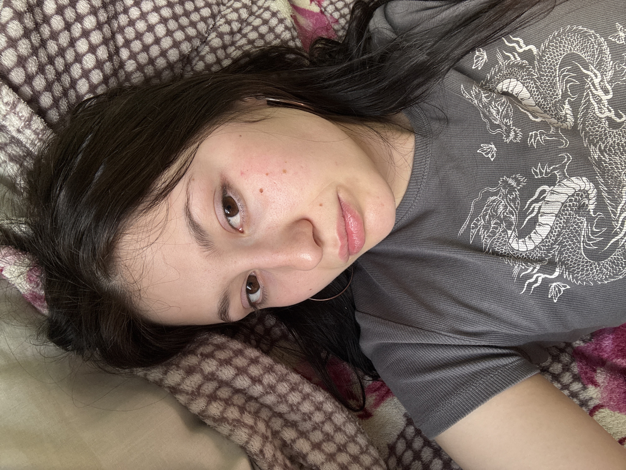 A woman is lying down on bedding with a patterned brown and white blanket in the foreground and a blanket with pink floral patterns in the background. She is wearing a grey t-shirt with a white dragon design. The woman has dark hair and is wearing earrings. No information about Фоки, Russia, is discernible from the image.