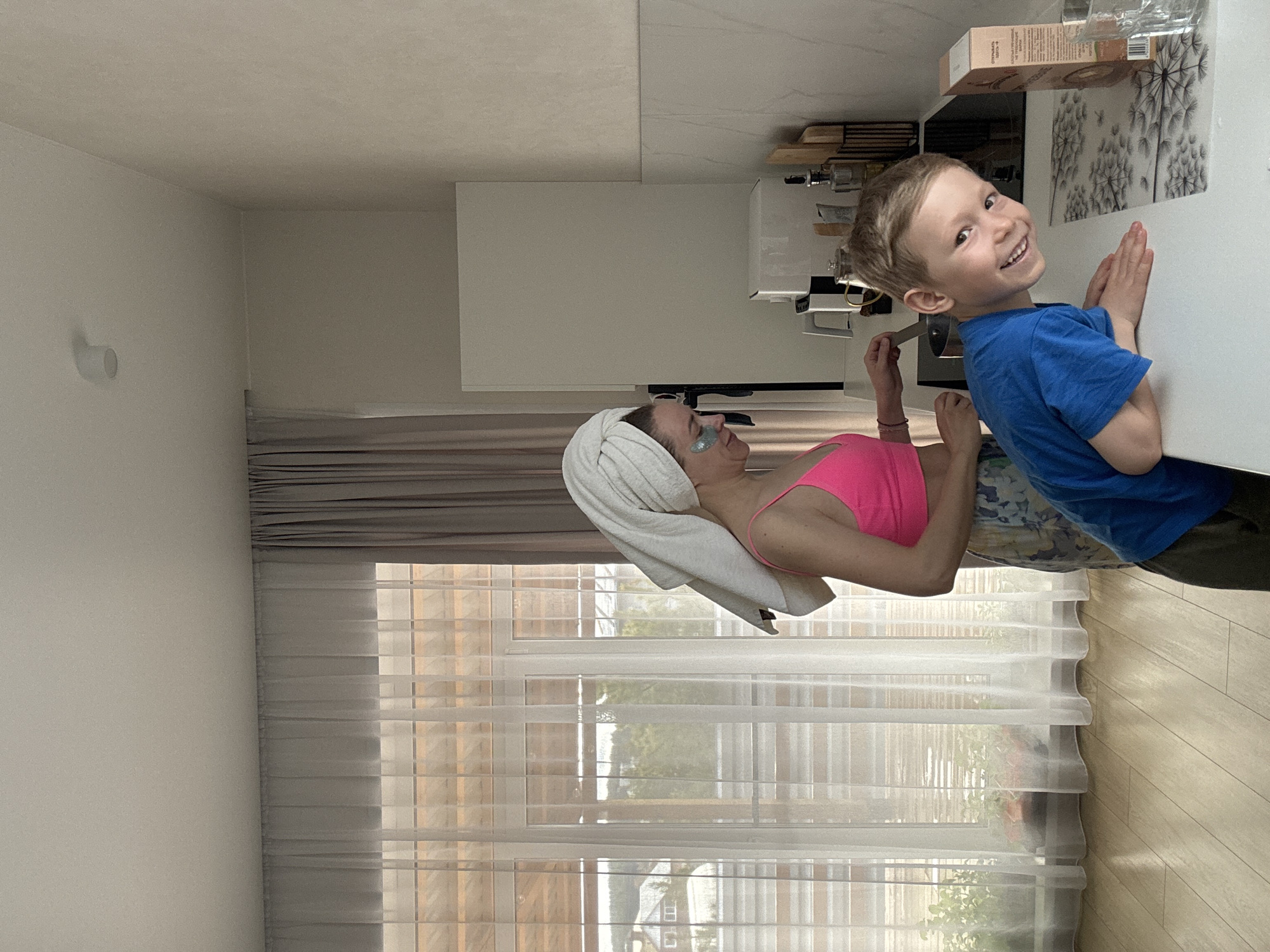 The scene appears to be inside a modern apartment. A woman with a towel wrapped around her head and eye patches is lying down. She is wearing a pink top. A young boy in a blue shirt is leaning on a white countertop next to her, smiling at the camera. There are kitchen appliances and other objects on the countertop. Light streams through a window behind the woman, revealing a view of what appears to be a residential neighborhood. It is not possible to confirm the location of Chaikovsky, Russia based on the visible objects in the image.