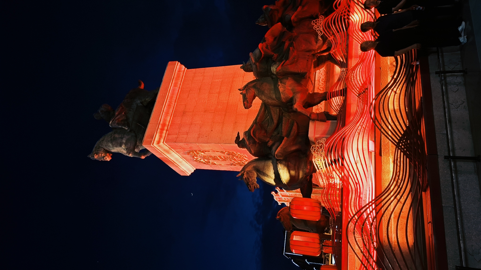 The image shows a statue in Yanta District, China, illuminated in red light against a dark blue sky. The statue depicts riders on horses mounted on a rectangular plinth. Adjacent to the statue are decorative red curved metal elements and traditional red lanterns. The image appears to be taken at night. Some people are visible in the upper right corner of the image.