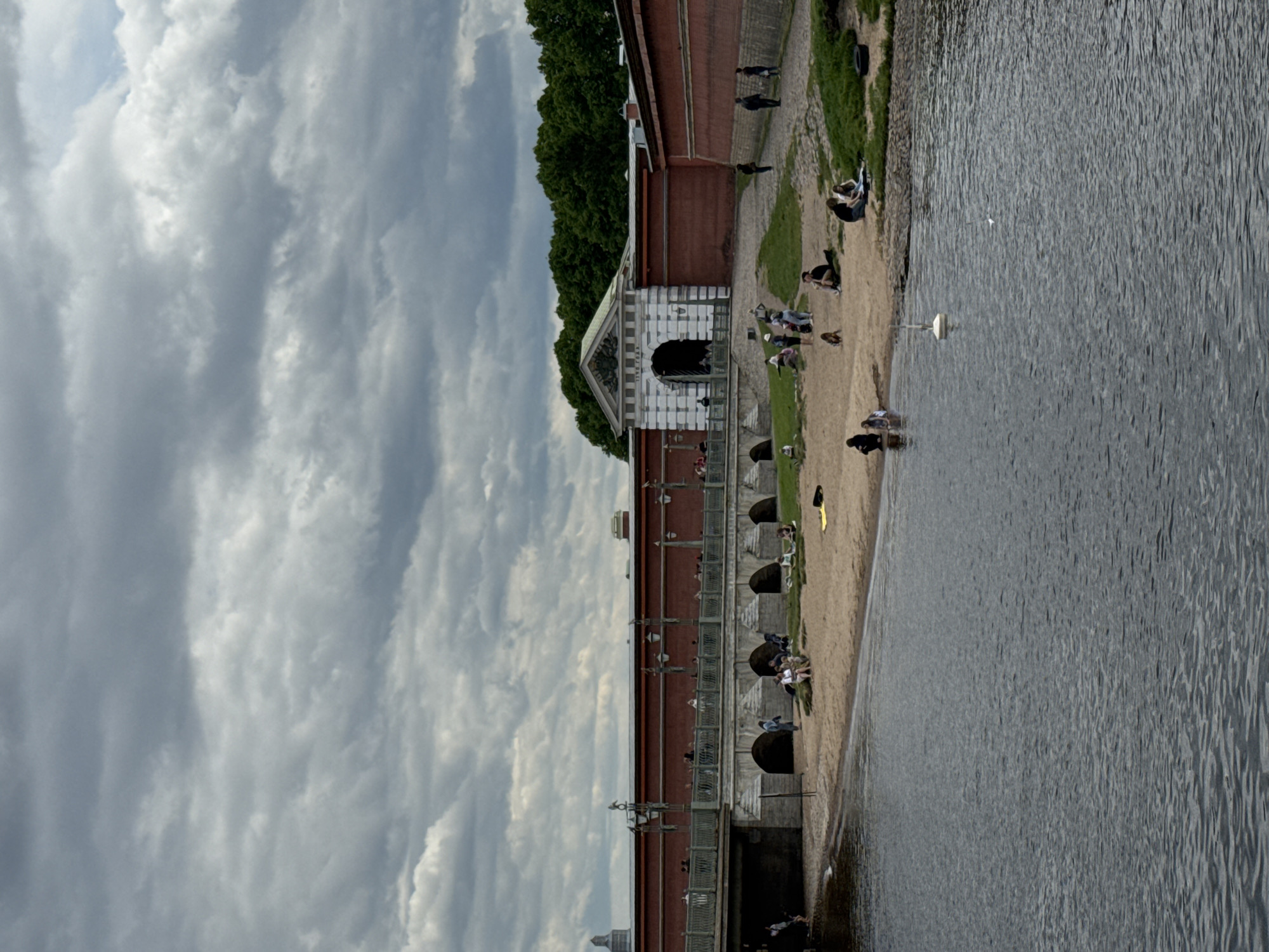 The image depicts a vibrant waterfront scene in Saint Petersburg, Russia, likely along the Neva River or a canal, with a historical red-brick fortress wall dominating the background. The sky is overcast with numerous grey clouds, indicating daytime.
In the foreground, a large body of water, likely a river, stretches across the frame, showing ripples on its surface. A small white buoy or float is visible in the water, and near it, a white bird, possibly a swan or goose, is swimming. To the left of the water, a sandy beach or bank extends, populated by numerous people enjoying leisure activities.
The main architectural feature is a long, imposing brick building or wall, possibly part of the Peter and Paul Fortress. It features multiple arched openings, some leading to dark interior spaces, and a distinctive entrance structure with white and grey striped detailing and a gabled roof. Green trees are visible behind and above parts of the red brick structure.
People are scattered along the sandy bank and the grassy areas adjacent to the buildings. Many individuals are sitting or lounging on the sand, some near the water's edge, possibly wading or sunbathing. Others are standing or walking along the pathways. One person is visible on the sandy bank with a yellow item nearby, possibly a towel or bag. The atmosphere appears relaxed and recreational. No clearly legible text is visible in the image.