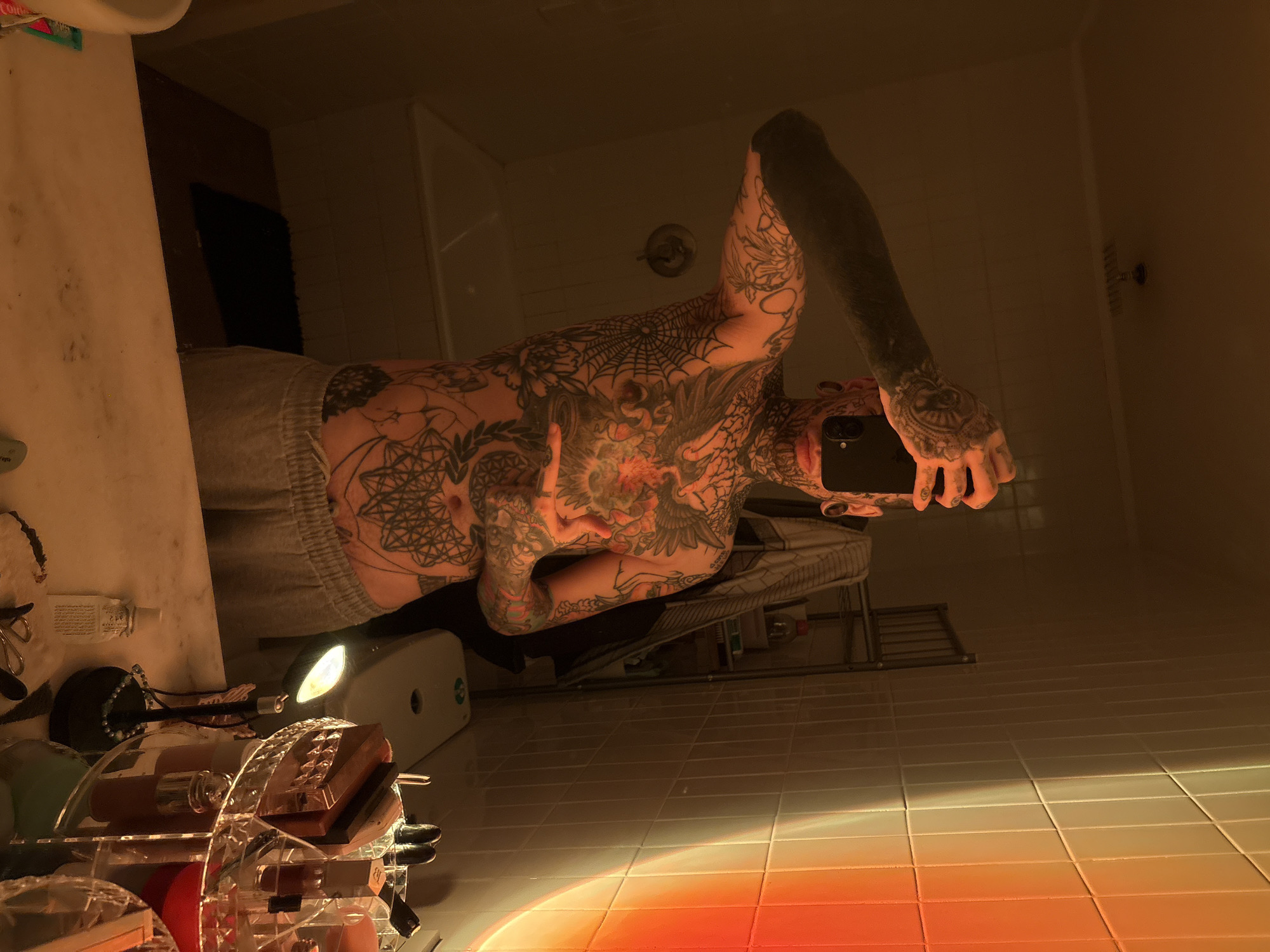 A person with extensive tattoos on their torso and arms stands in a bathroom taking a selfie in a mirror. The person is wearing grey sweatpants. The bathroom has tiled walls and floors, with a shower visible in the background. A sink and vanity area are present to the side, with various cosmetic items on the countertop. The lighting is warm and gives the tiles on the floor a gradient from light to dark orange. There is no indication of the city of Buenos Aires, Argentina, or any specific location.