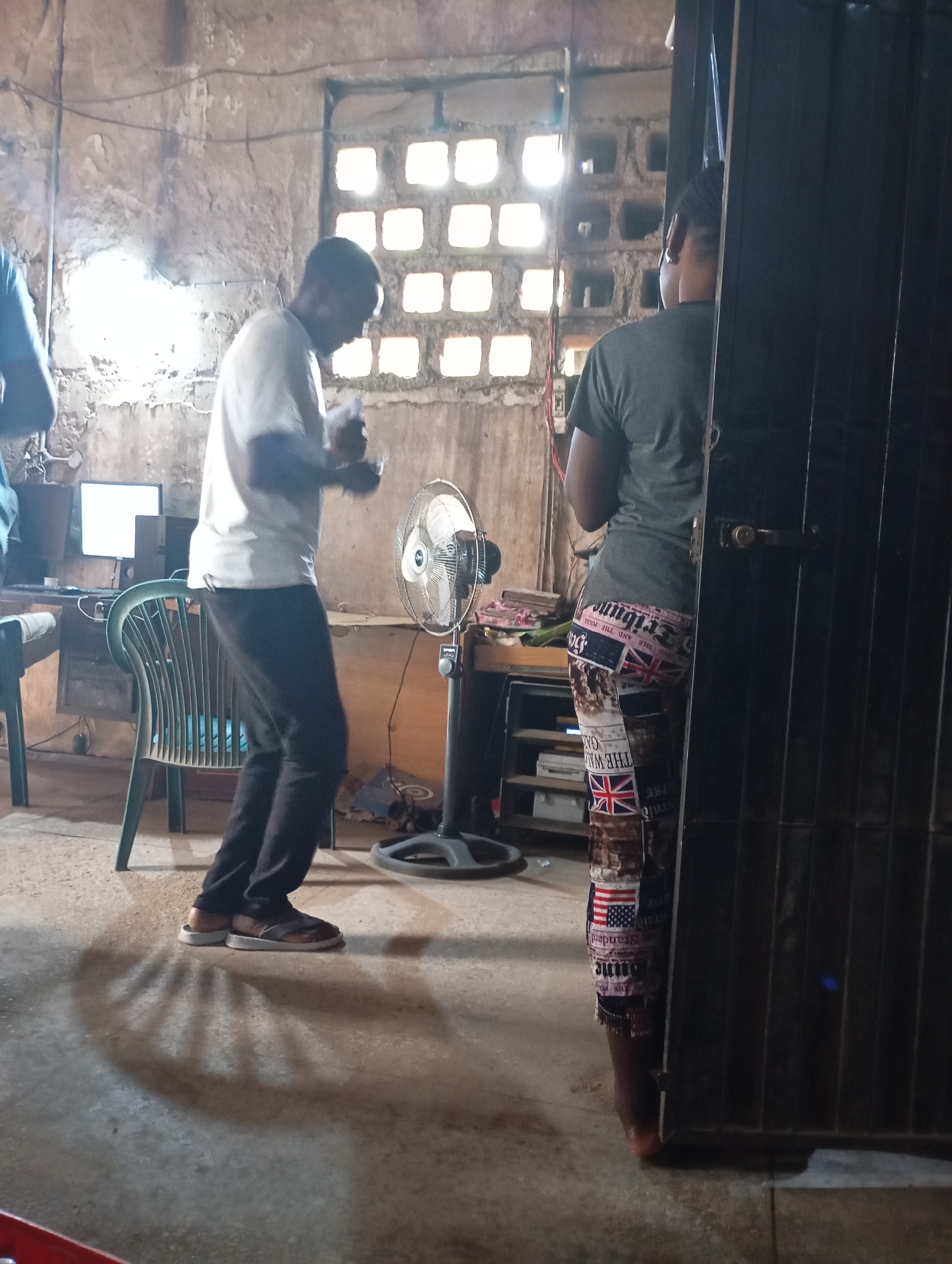 In Shendam, Nigeria, a man wearing a white t-shirt and dark pants stands with his back to a wall, holding a piece of paper. He is barefoot. To his right, a woman in a grey t-shirt and patterned leggings stands facing away from the camera, with her left side visible. She appears to be standing by a dark doorway. In the background, a pedestal fan is positioned between the two individuals, and a computer monitor and chair are visible to the left of the man. The room is sparsely furnished with rough, textured walls and a window with square openings. The floor is concrete, and shadows are cast on it from the objects and people in the room.