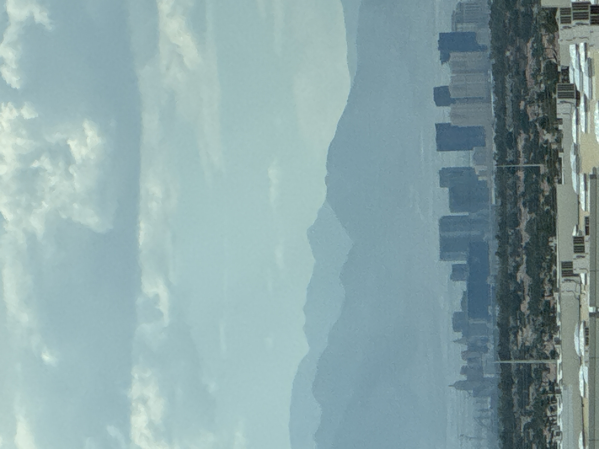 This image depicts a hazy, distant view of the Las Vegas Strip skyline, viewed from the city of Henderson, United States of America. The foreground features the rooftops of buildings, possibly commercial or industrial, with visible air conditioning units and some white coverings. Beyond these rooftops, a dense green area, likely vegetation or trees, separates the foreground from the city. The middle ground is dominated by the cluster of high-rise buildings that constitute the Las Vegas Strip. These buildings appear somewhat softened by the atmospheric haze, with various architectural styles visible, including some with distinct shapes and glass facades. In the background, a series of layered mountain ranges fade into the distance, their forms becoming progressively fainter and bluer due to atmospheric perspective. The sky above is filled with soft, diffused clouds, contributing to the overall muted and hazy quality of the scene. The lighting suggests it is daytime, but the haze obscures sharp details and the specific time of day. There are no visible people or distinct actions occurring.