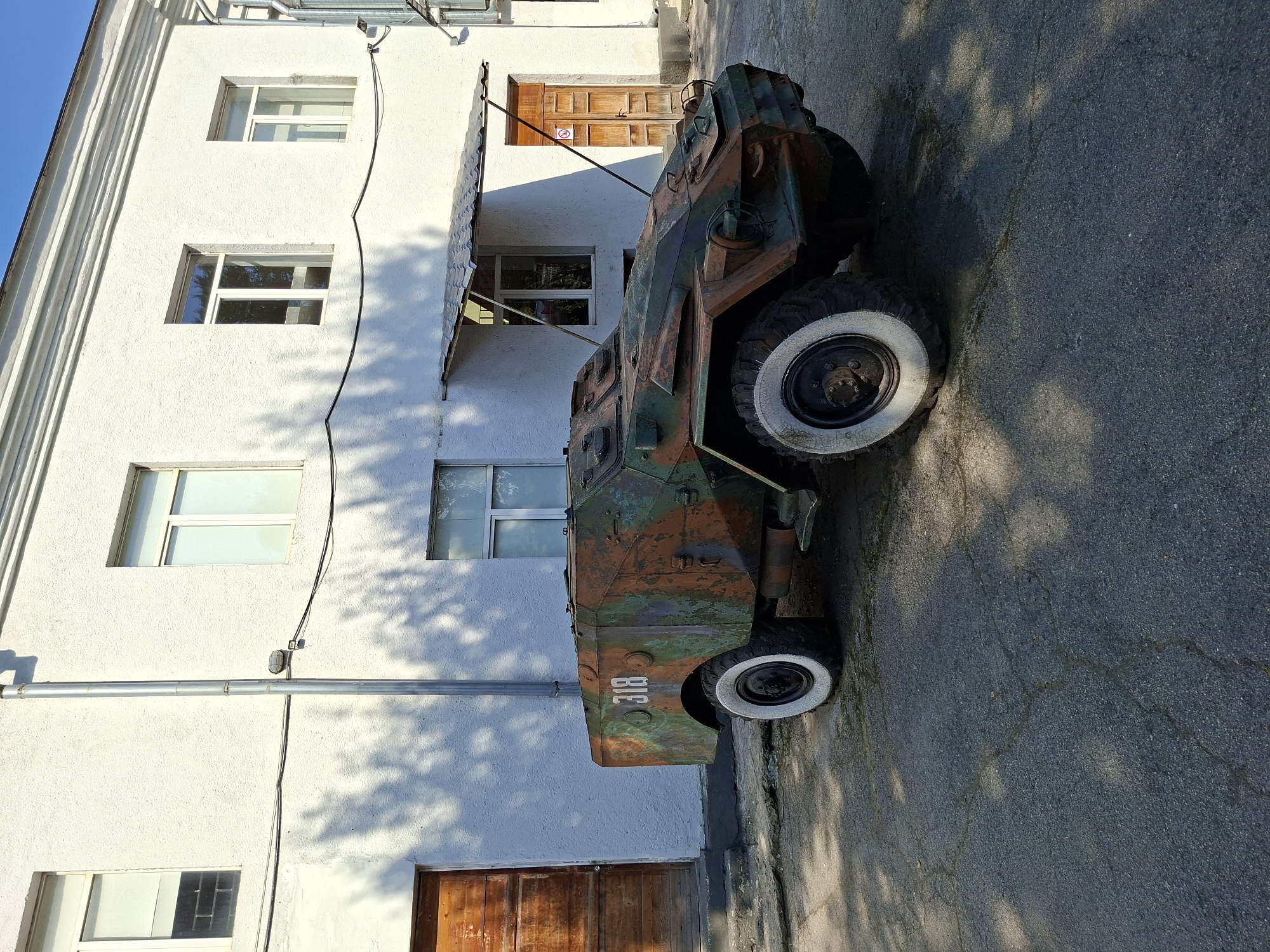 The image depicts a heavily weathered, camouflage-painted armored vehicle parked on asphalt outside a white, textured building. The vehicle, which appears to be a vintage military vehicle, has the number "318" painted in white on its side. It has large, rugged tires, with at least one of them having a white rim. The building behind the vehicle has several windows, some with blinds, and two wooden doors. A metal drainpipe runs vertically along the exterior wall. The ground shows signs of cracking and wear. Shadows of tree branches are cast across the building and the ground, indicating it is daytime and likely sunny. This scene is consistent with the grounds of a museum or historical site. Based on visual cues and common knowledge of such historical displays, this scene could be located at the Military Museum in Chișinău, Moldova, known for exhibiting military equipment. No people are visible in the image.