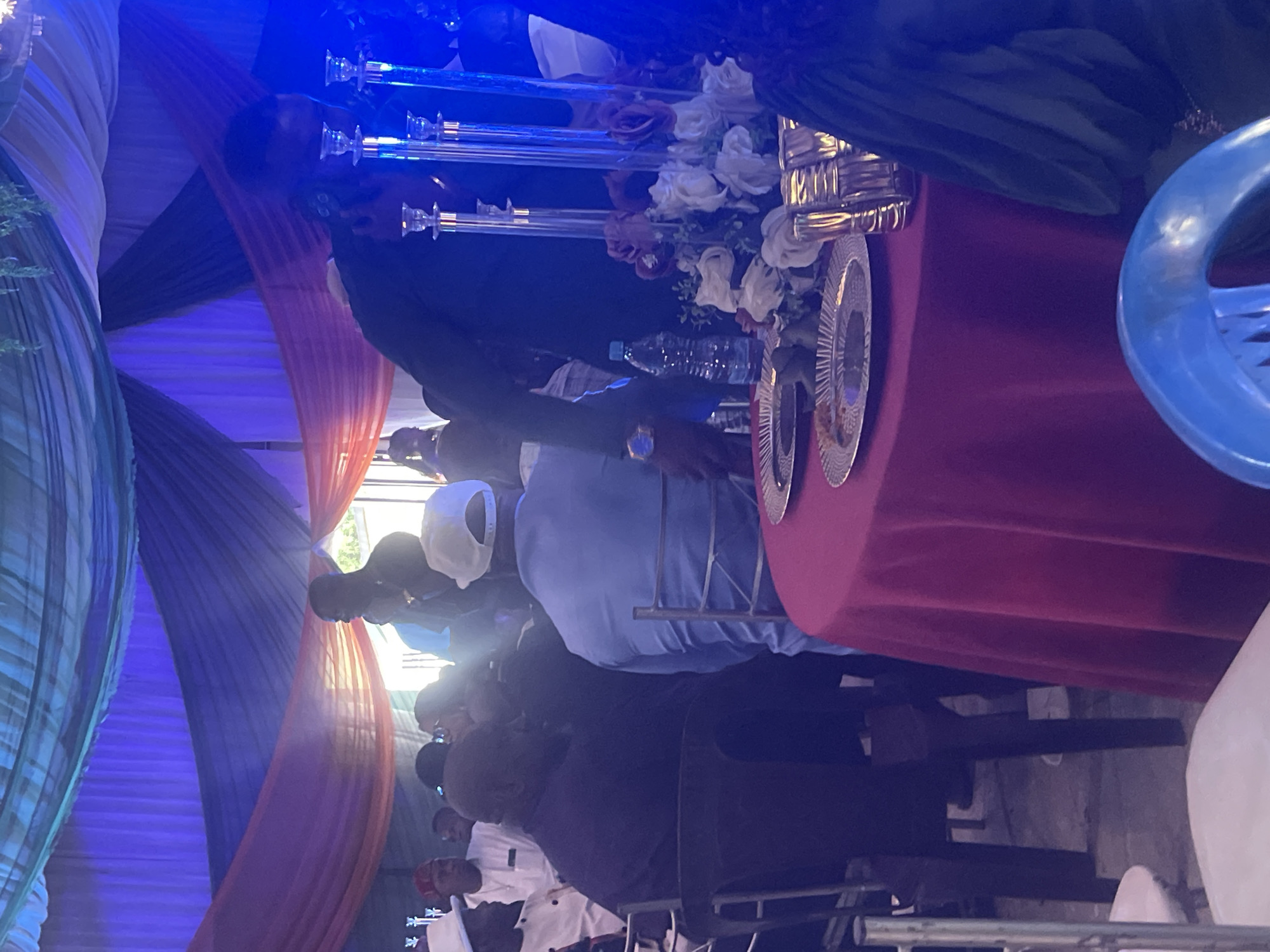 The image shows an event in progress, possibly a wedding or celebration, taking place in Jos, Nigeria. Attendees are seated, including men in suits and casual attire. A red-draped table is the focal point, adorned with floral arrangements, decorative plates, and tall candle holders. Behind the table, draped fabric creates a backdrop with blue and peach tones. Blue lighting enhances the environment.