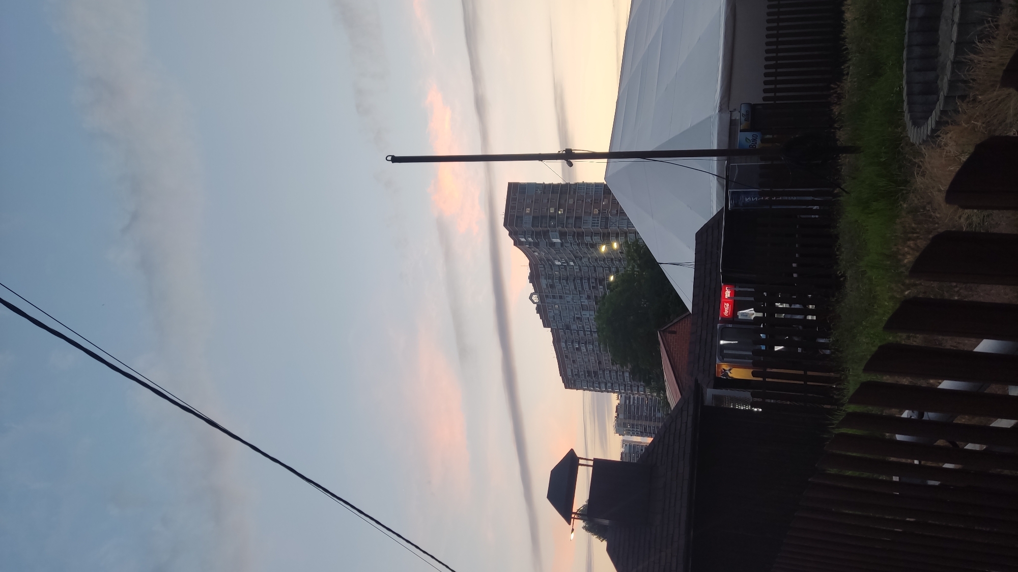 The image depicts a cityscape at dusk, with a prominent, curved residential building dominating the background. The sky is a soft gradient of blue and pink, with elongated clouds streaking across it. In the foreground, a dark wooden fence runs across the bottom of the frame. To the left, a dark, angular structure with a pitched roof and a small, pointed upper section is visible. Adjacent to this, a larger, white, tent-like structure is partially visible on the right. Between the foreground structures and the tall building, several smaller buildings and trees are present. A Coca-Cola logo is illuminated on one of the smaller buildings. There are no people visible in the image. The overall scene suggests an urban environment, possibly a recreational area or event space, set against the backdrop of apartment buildings as daylight fades.