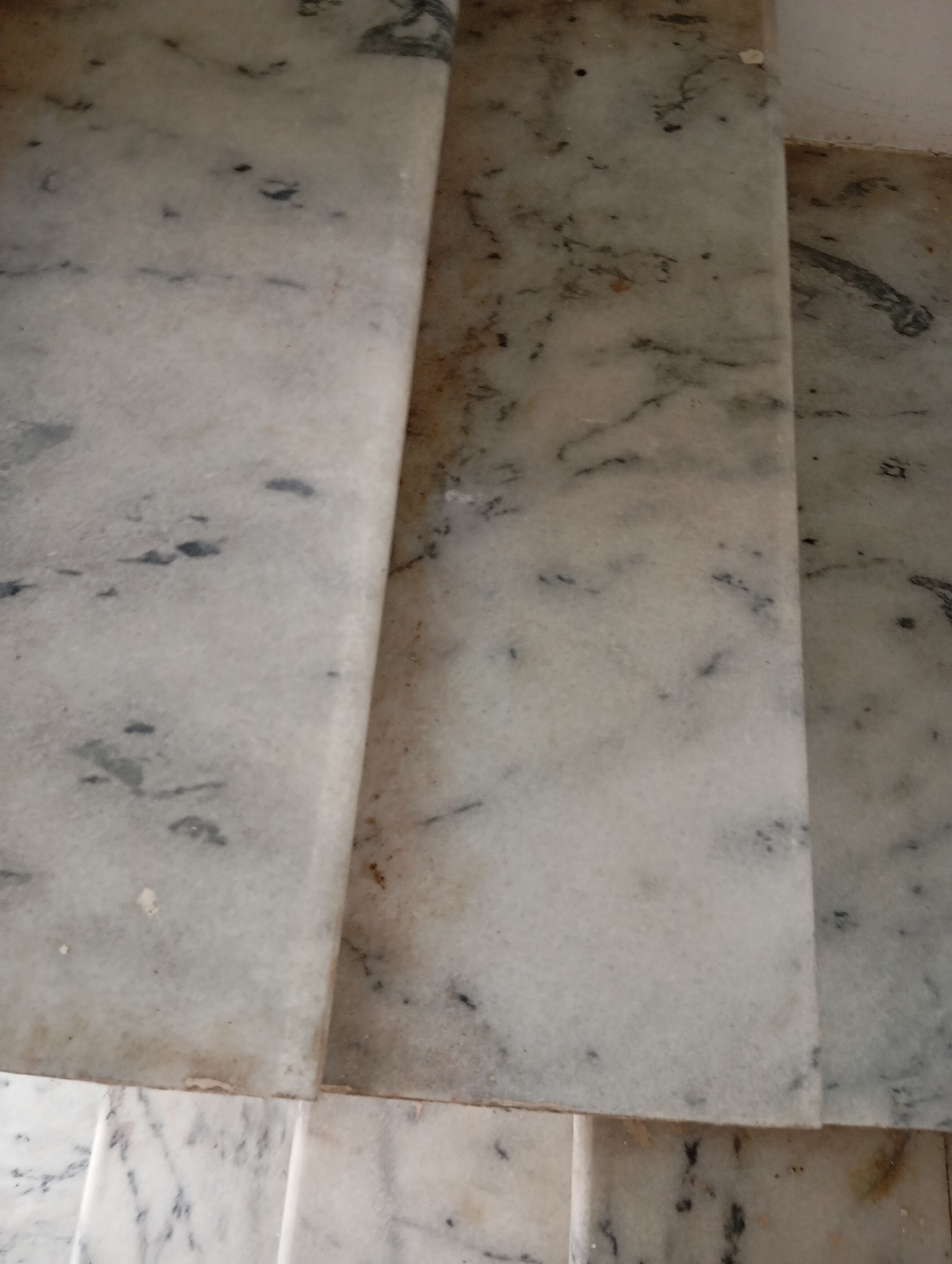The image shows a close-up, overhead view of marble steps. The marble is light-colored with dark gray veining. The steps appear to be part of a staircase, with the top step partially visible in the foreground and extending to the right. Below the top step, another step is visible, appearing slightly narrower. The surface of the marble is textured and shows some signs of wear, with small dark specks and lighter discolored areas scattered across its surface. The lighting is even, casting no distinct shadows. The environment is indoors, indicated by the smooth, finished surfaces of the marble. No people or other objects are present in the frame.