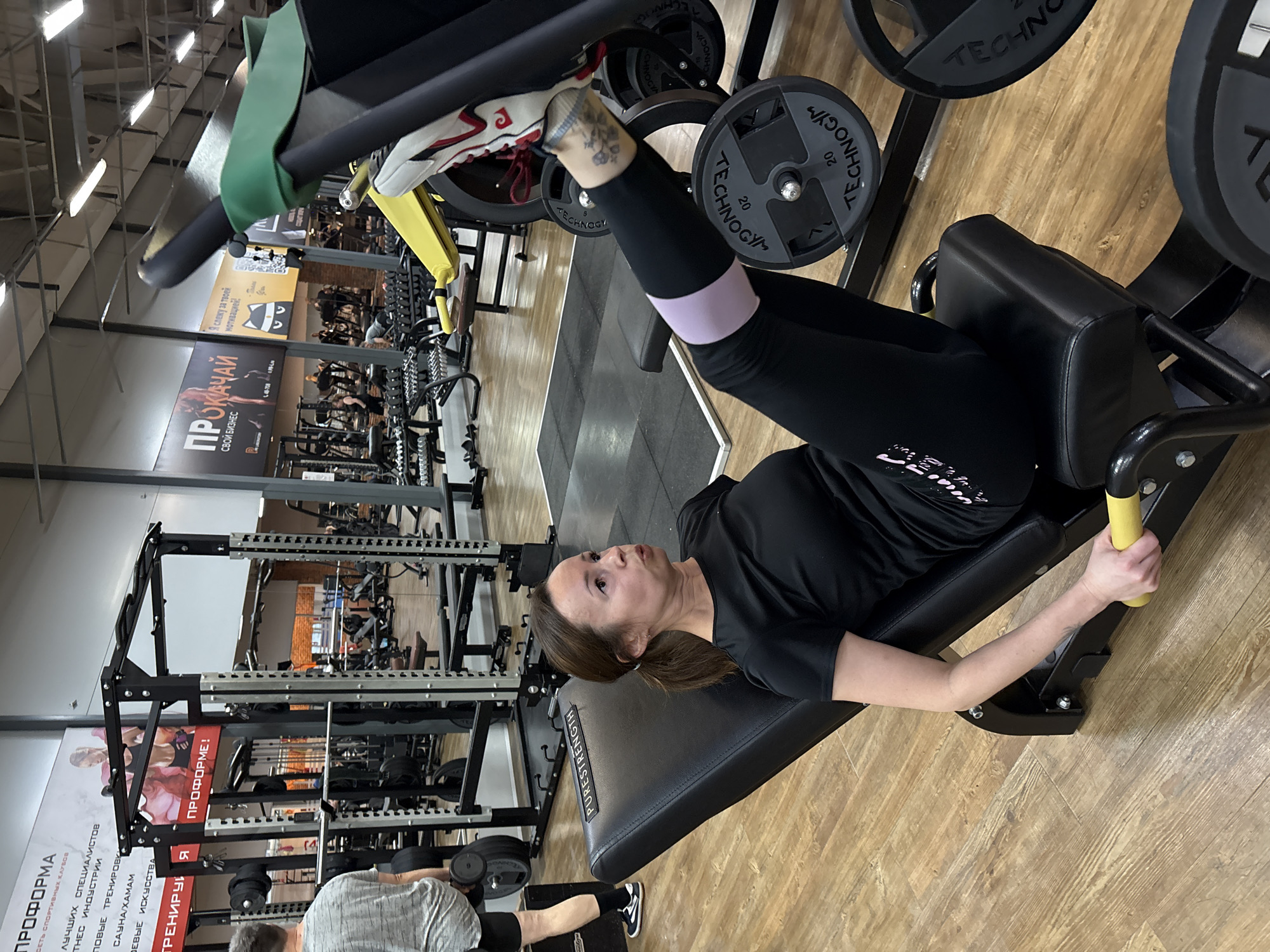Here is a detailed description of the image:
**Scene:** The image appears to be taken inside a gym in Chaikovsky, Russia, as evidenced by the Cyrillic writing on the wall and the various exercise machines in the background. The environment includes weight machines, dumbbells, and motivational posters on the walls. The floor is light-colored wood.
**Subject:** The primary subject is a woman performing a leg press exercise. She is lying on the leg press machine with her legs positioned to push the weighted platform. She is wearing black leggings with a light purple band around her upper thigh, a black t-shirt, and athletic shoes. There is a tattoo on her ankle. Her ponytail is visible.
**Objects:**
- A leg press machine with black weight plates.
- A weight rack is visible in the background.
- Dumbbells on the floor.
- Gym posters and motivational signs written in Cyrillic.
- Black equipment
**People:** Another individual is visible in the lower left corner, using dumbbells.
